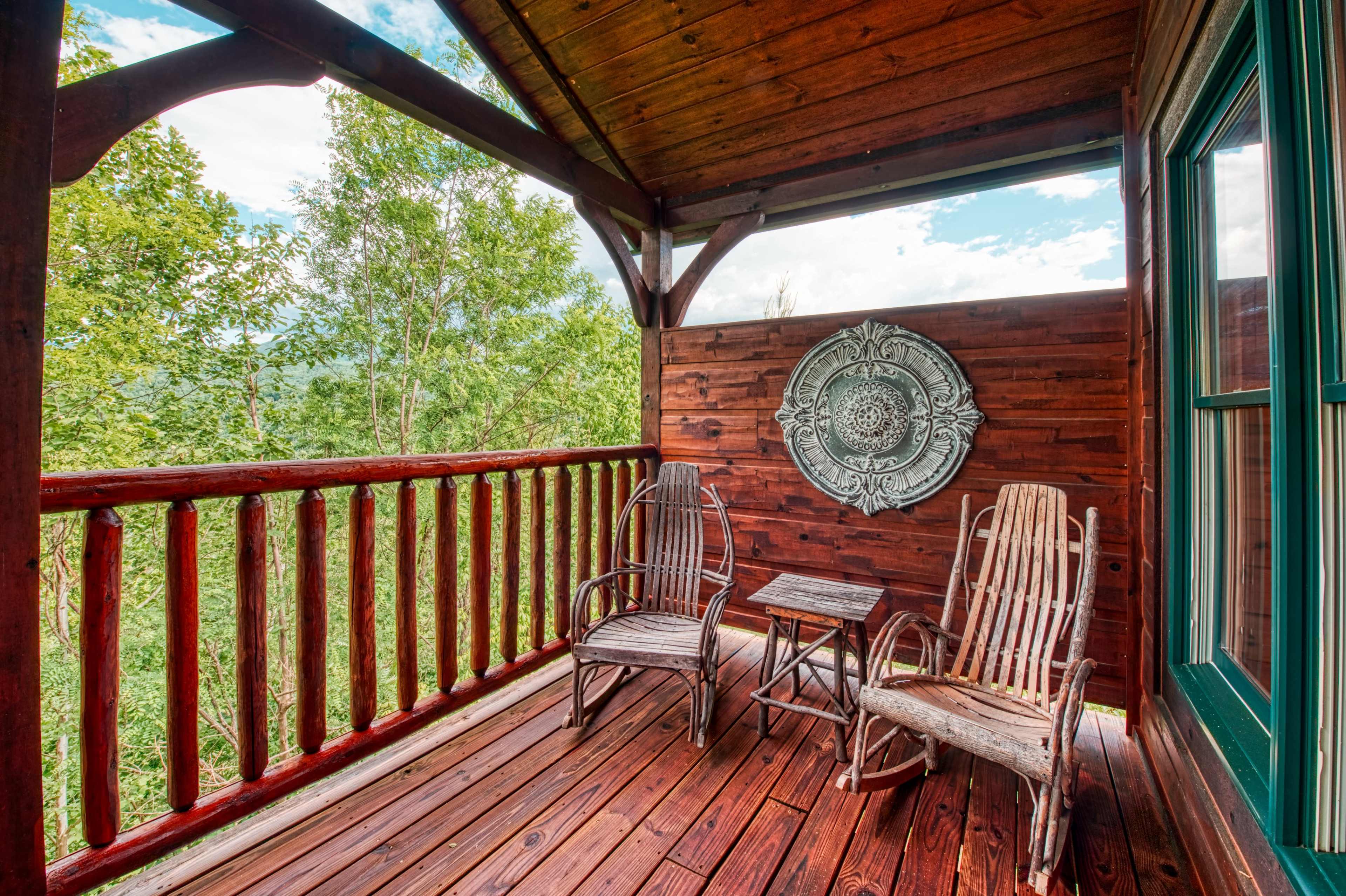 A wooden balcony with two rocking chairs and a small table, surrounded by trees and mountains in the background.