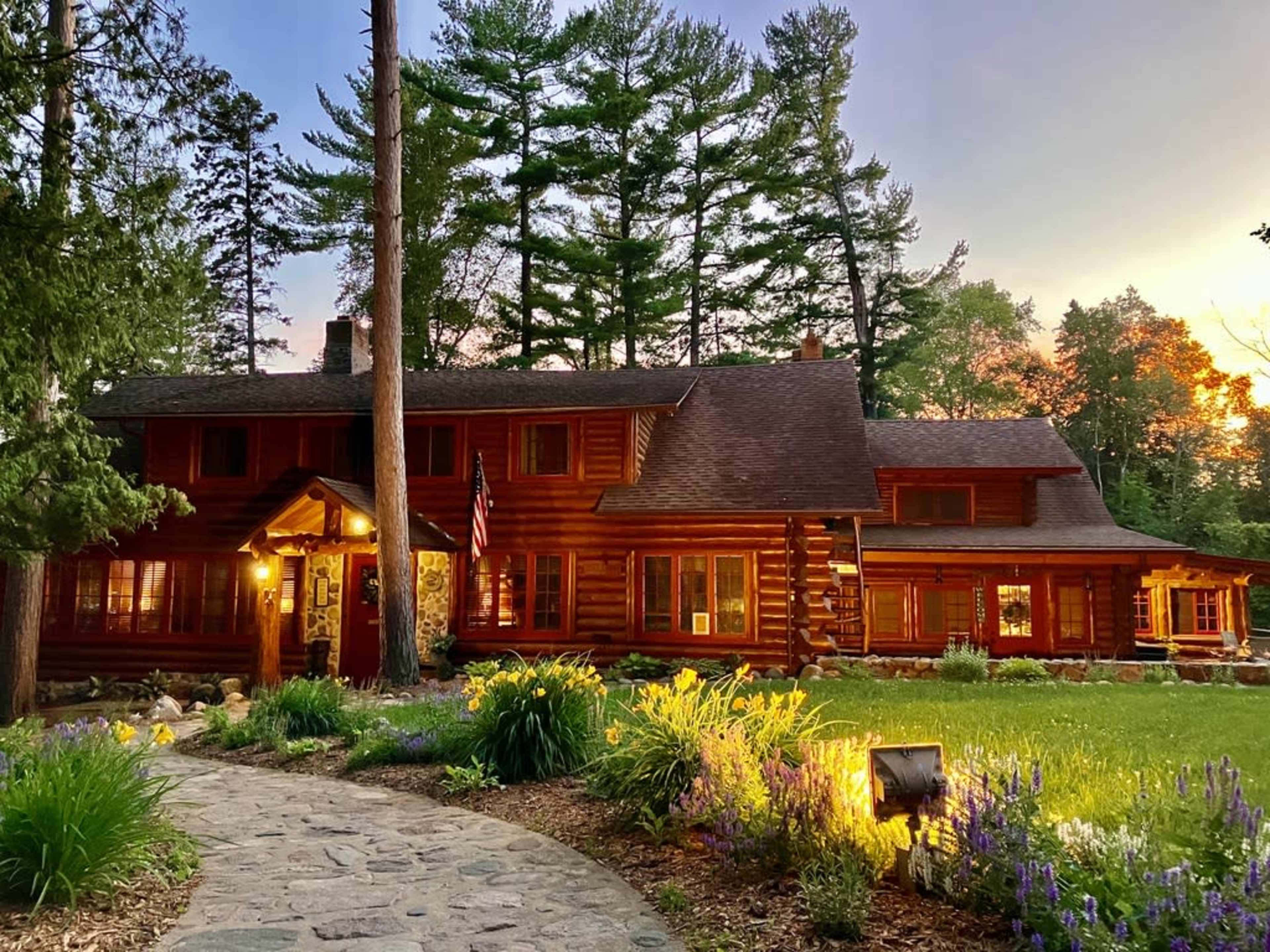 A large log cabin with a warm exterior is surrounded by trees and colorful flowers, with a stone pathway leading to its entrance.