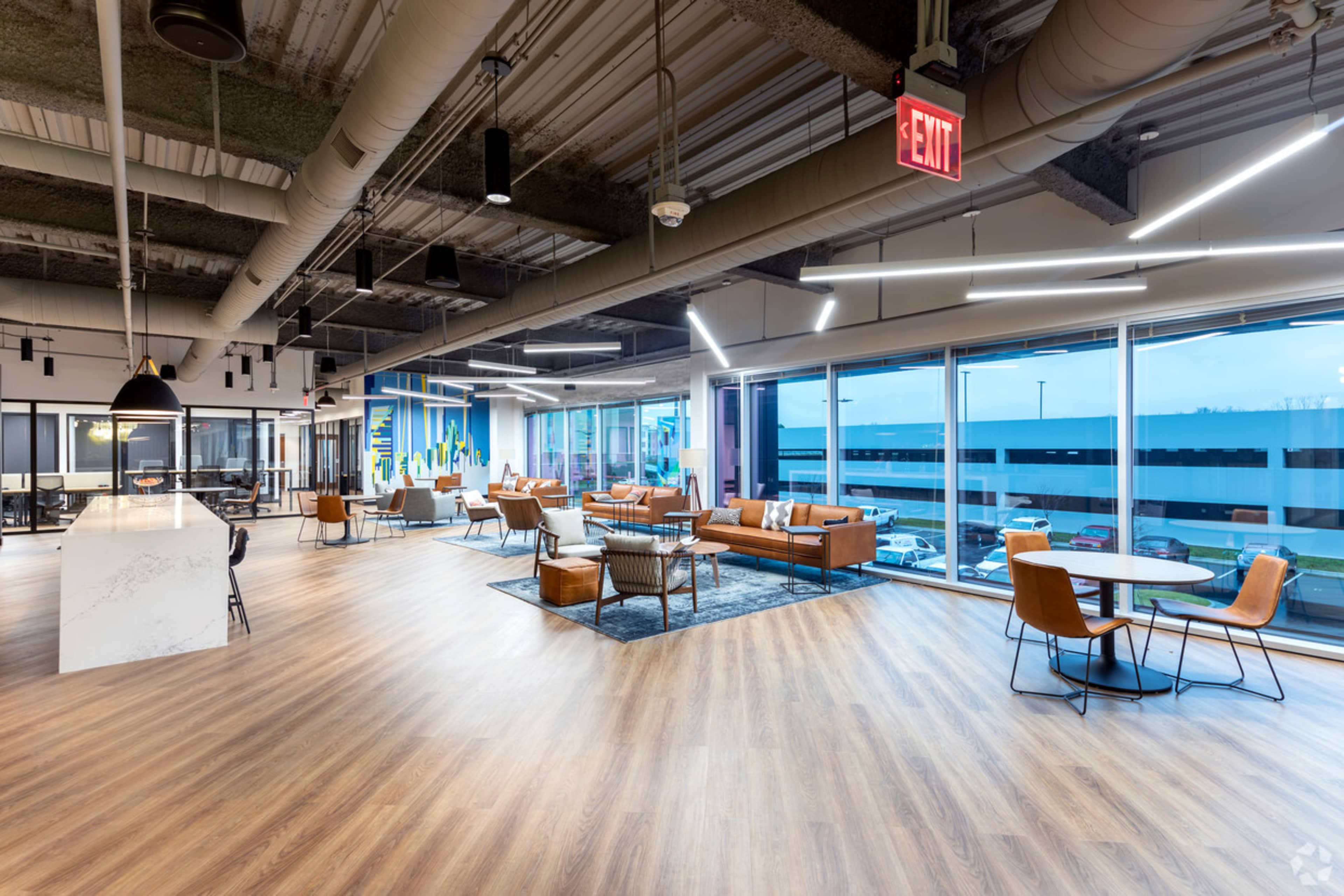 A modern office interior featuring leather seating, wooden flooring, large windows, and a mix of work and relaxation areas.