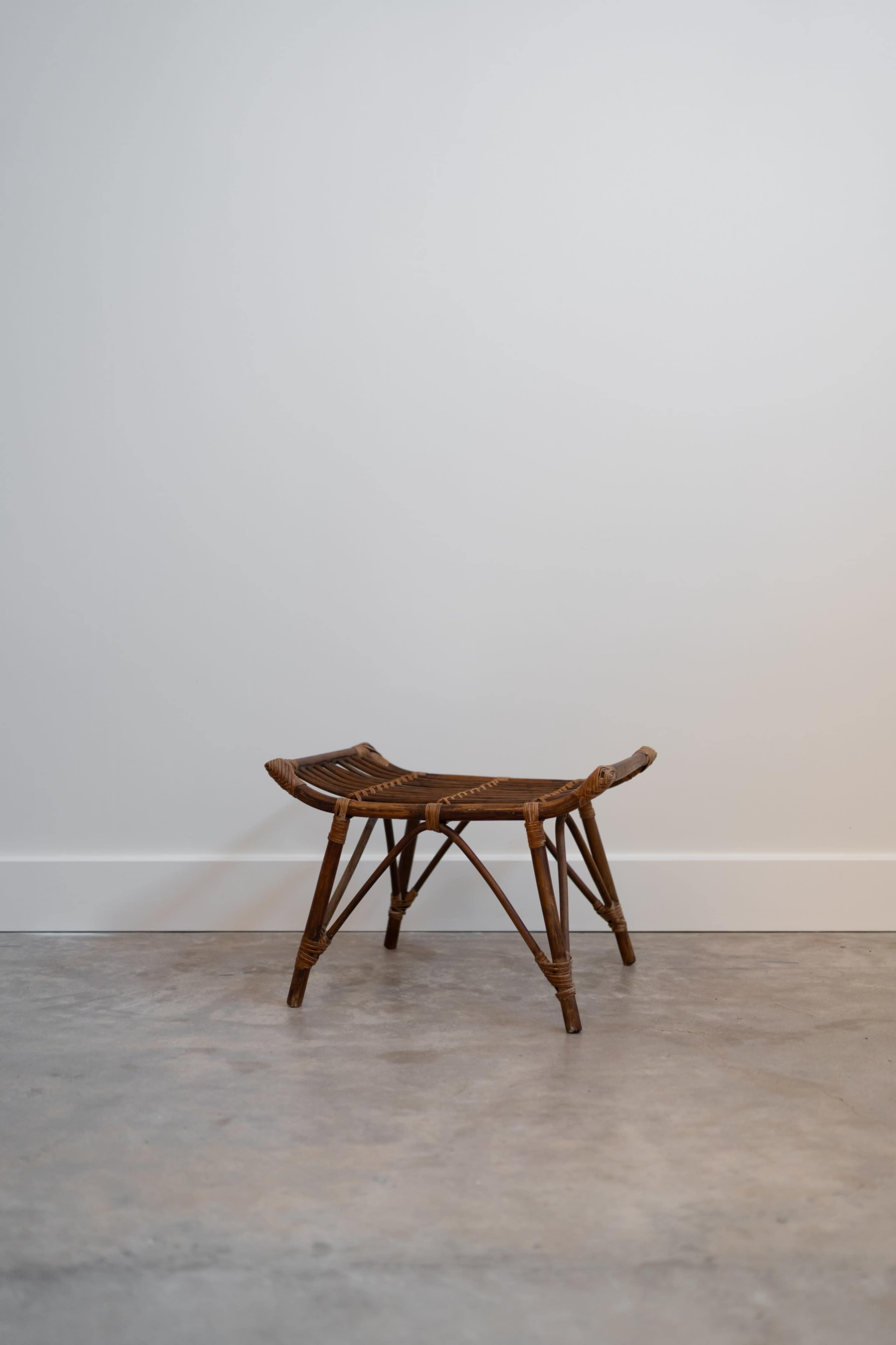 A wooden stool with a simple design sits against a plain white wall on a concrete floor.