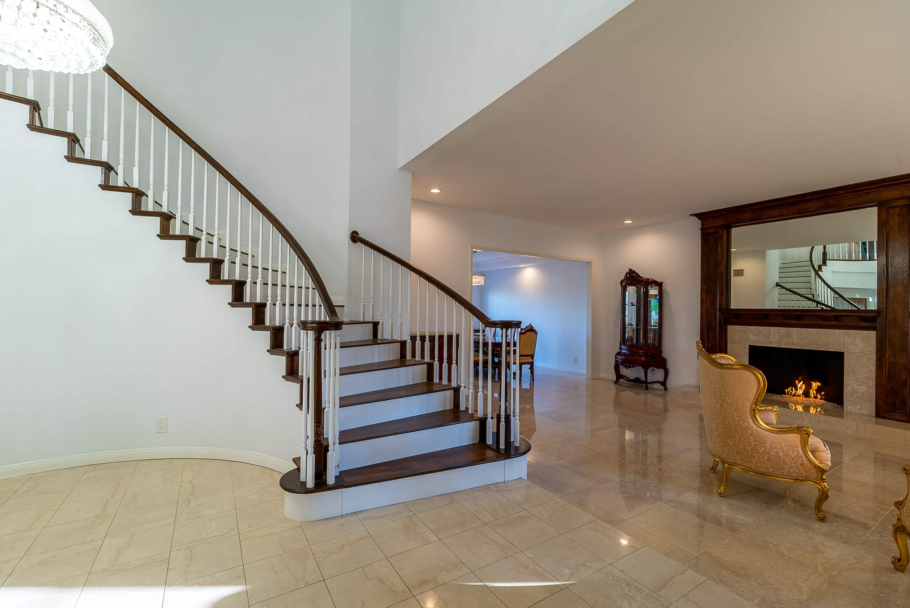 A curved staircase with white railings is the focal point of a spacious foyer featuring a gleaming marble floor and a cozy seating area beside a fireplace.