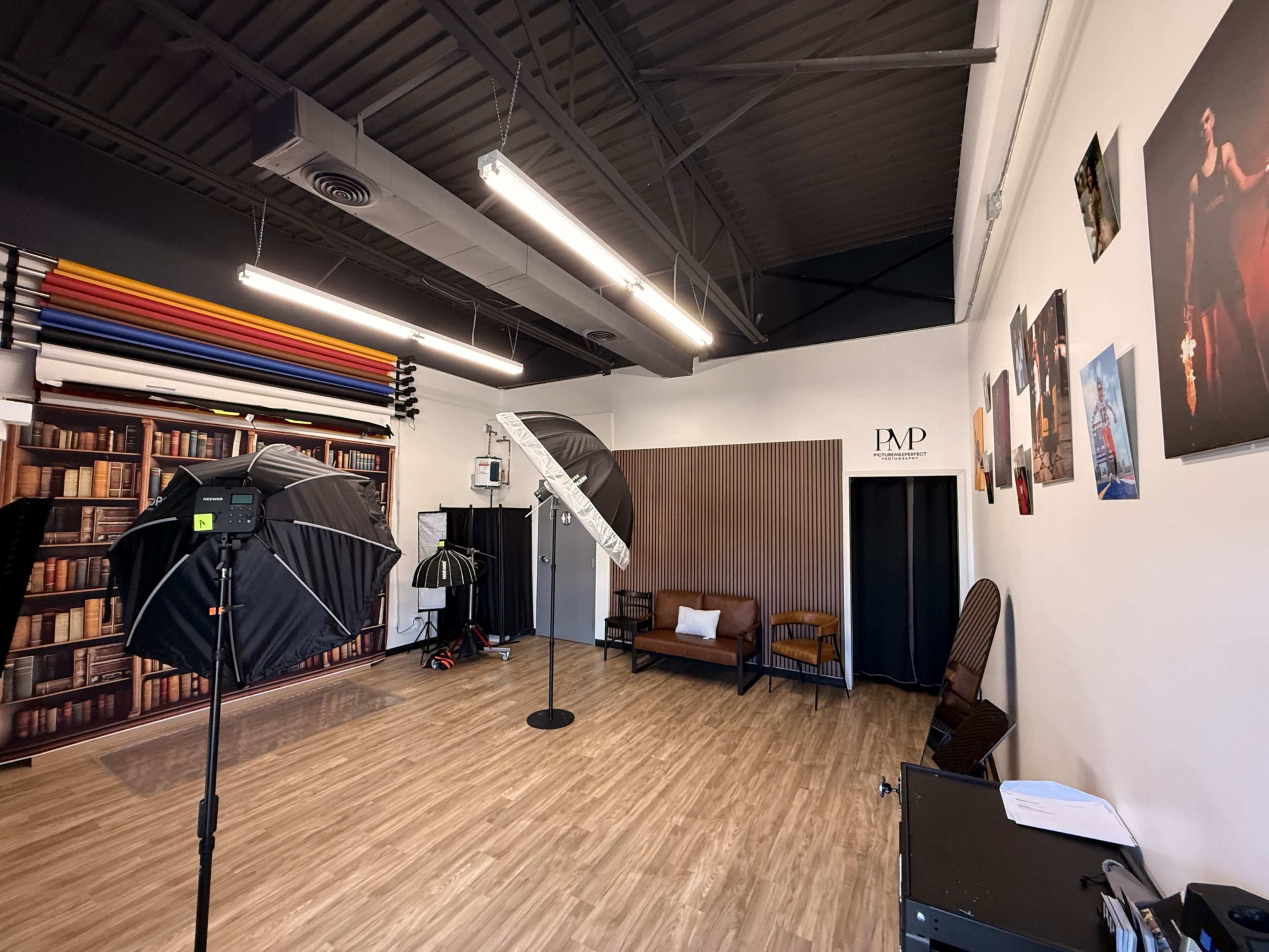 The image shows a photography studio featuring a backdrop of colorful rolls, two light setups, a brown couch, and various framed photos on the walls.