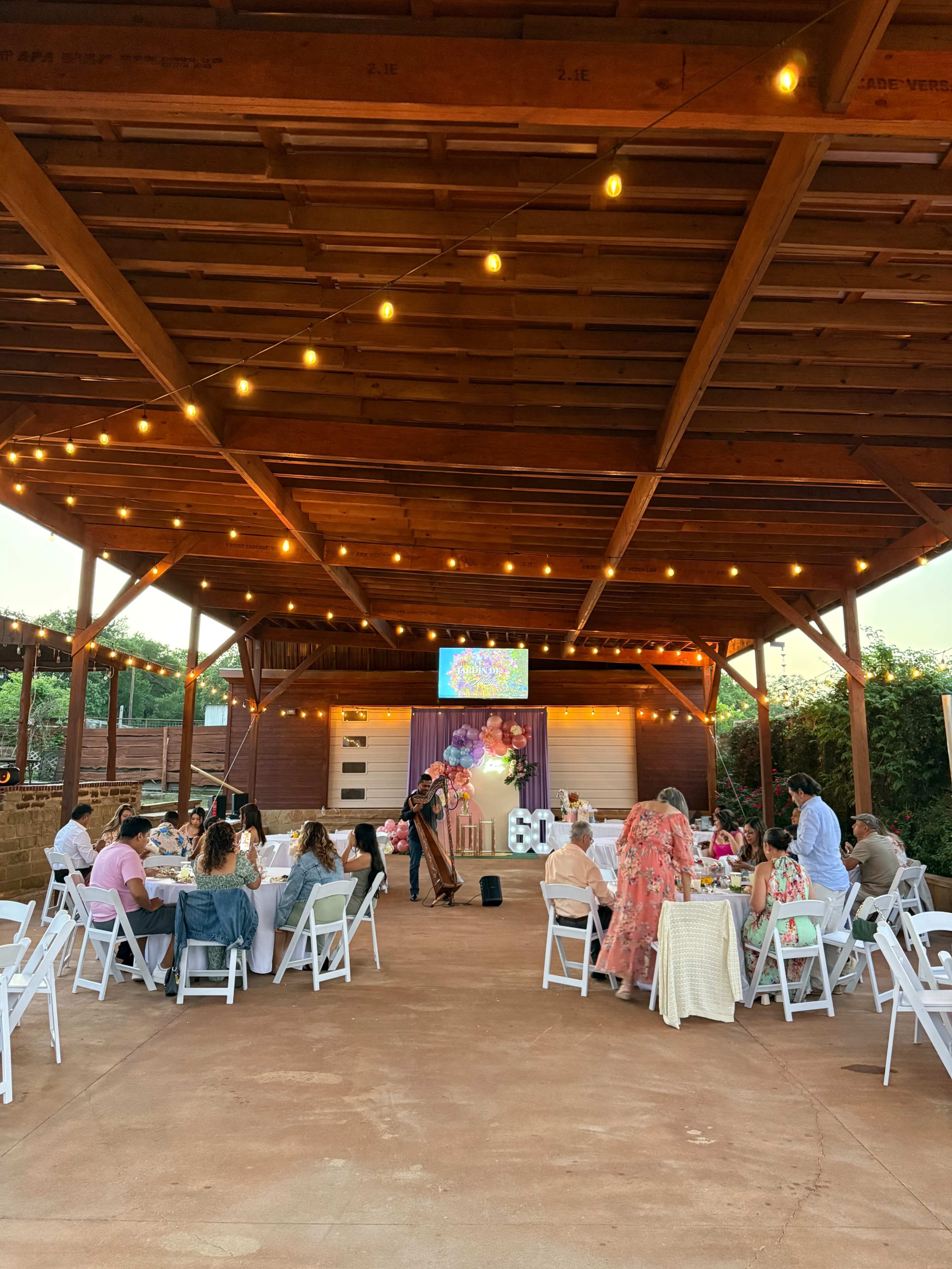 A group of people sits at tables under a wooden pavilion with string lights, while a musician performs on a small stage.