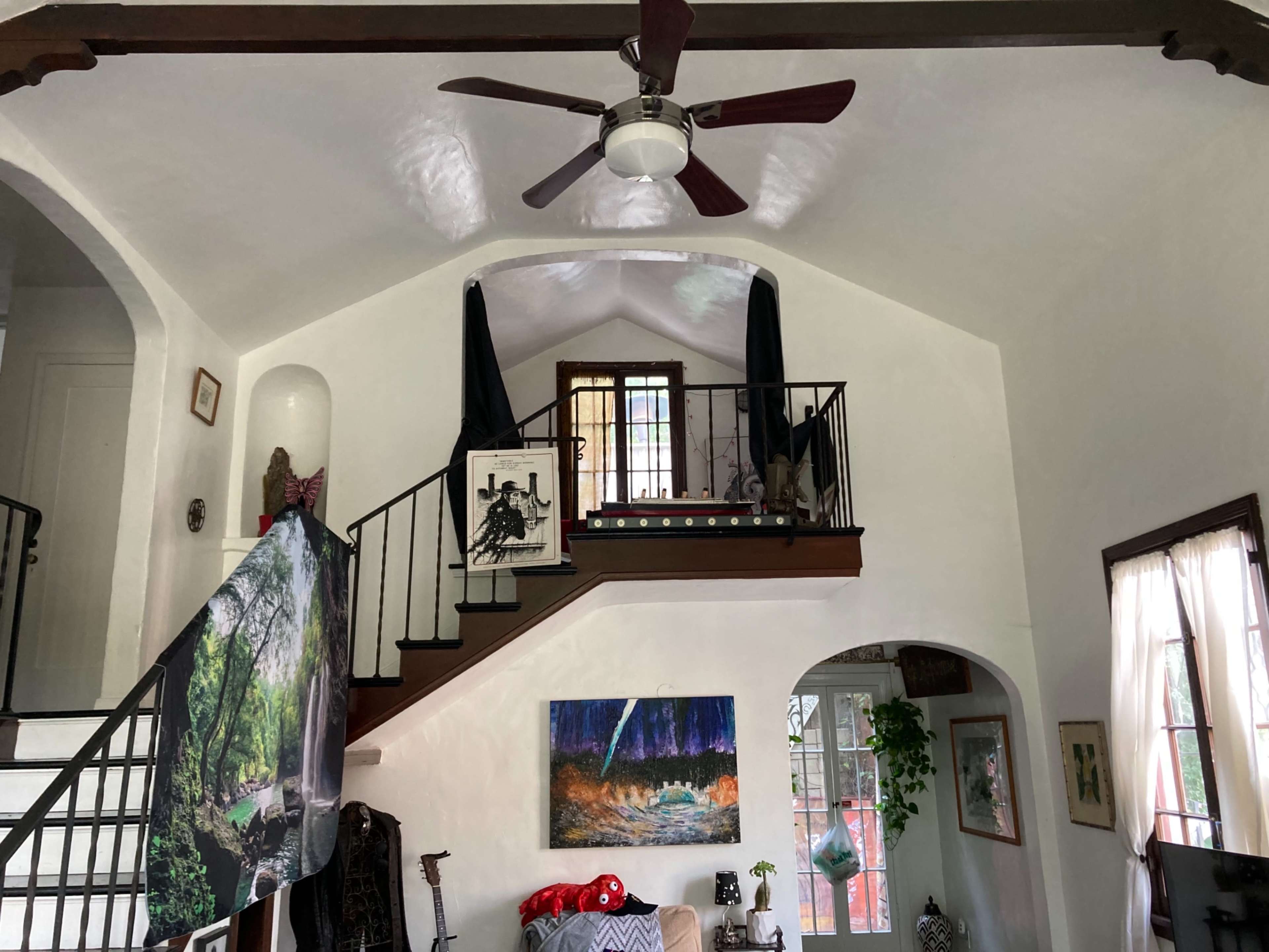 A living room with a staircase leading to a small upper landing, featuring artwork on the walls and a ceiling fan.