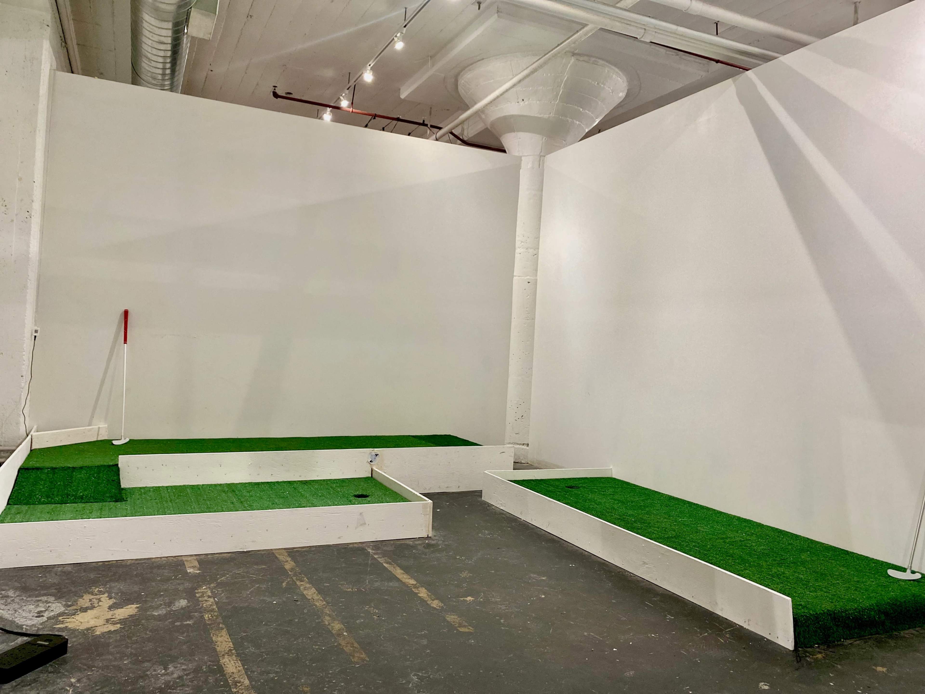 The image shows an indoor putting green setup with two tiers of artificial turf and a white wall in the background.