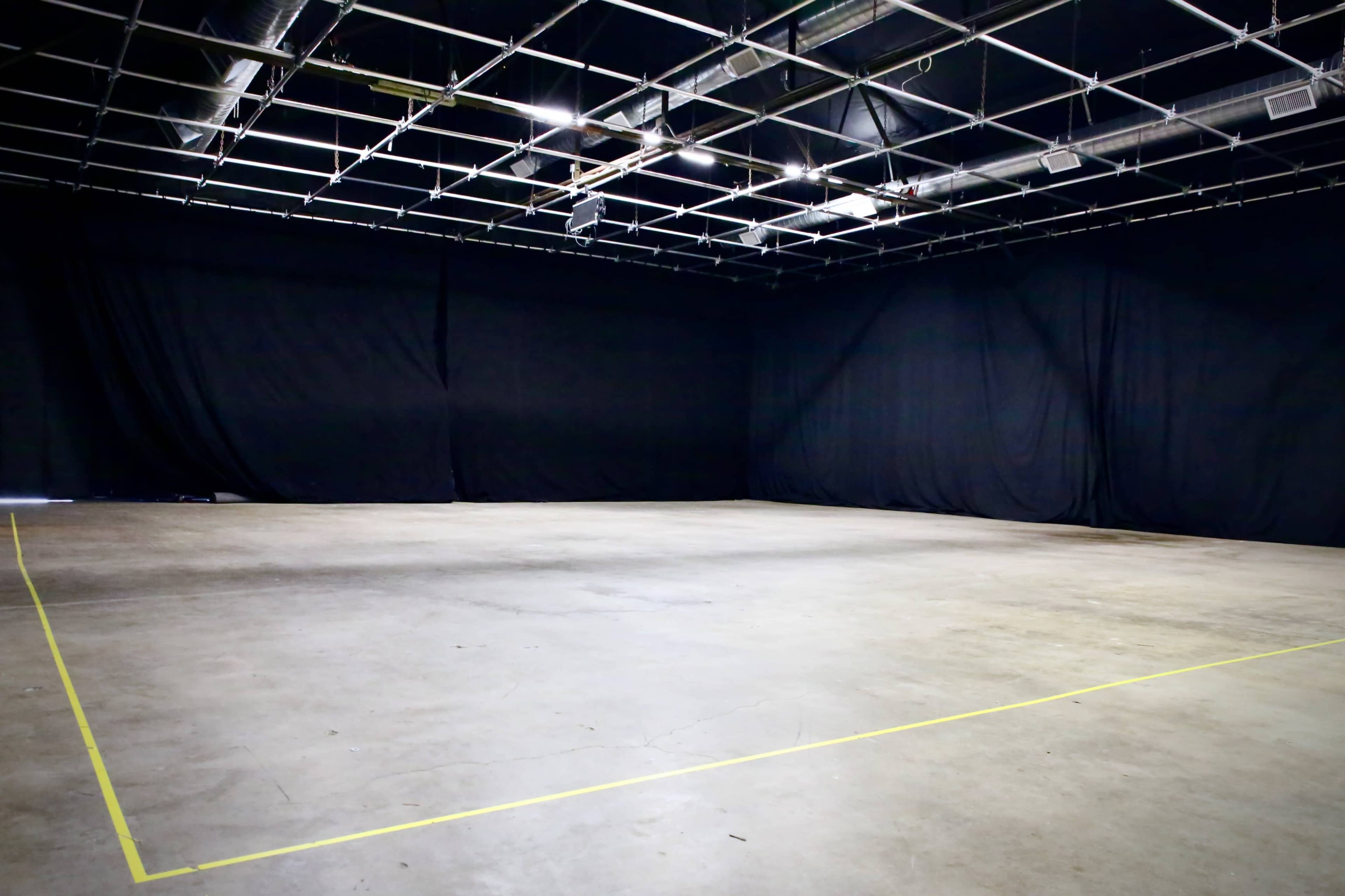 A large, empty studio space with black curtains and a concrete floor, marked by yellow tape outlining a rectangular area.