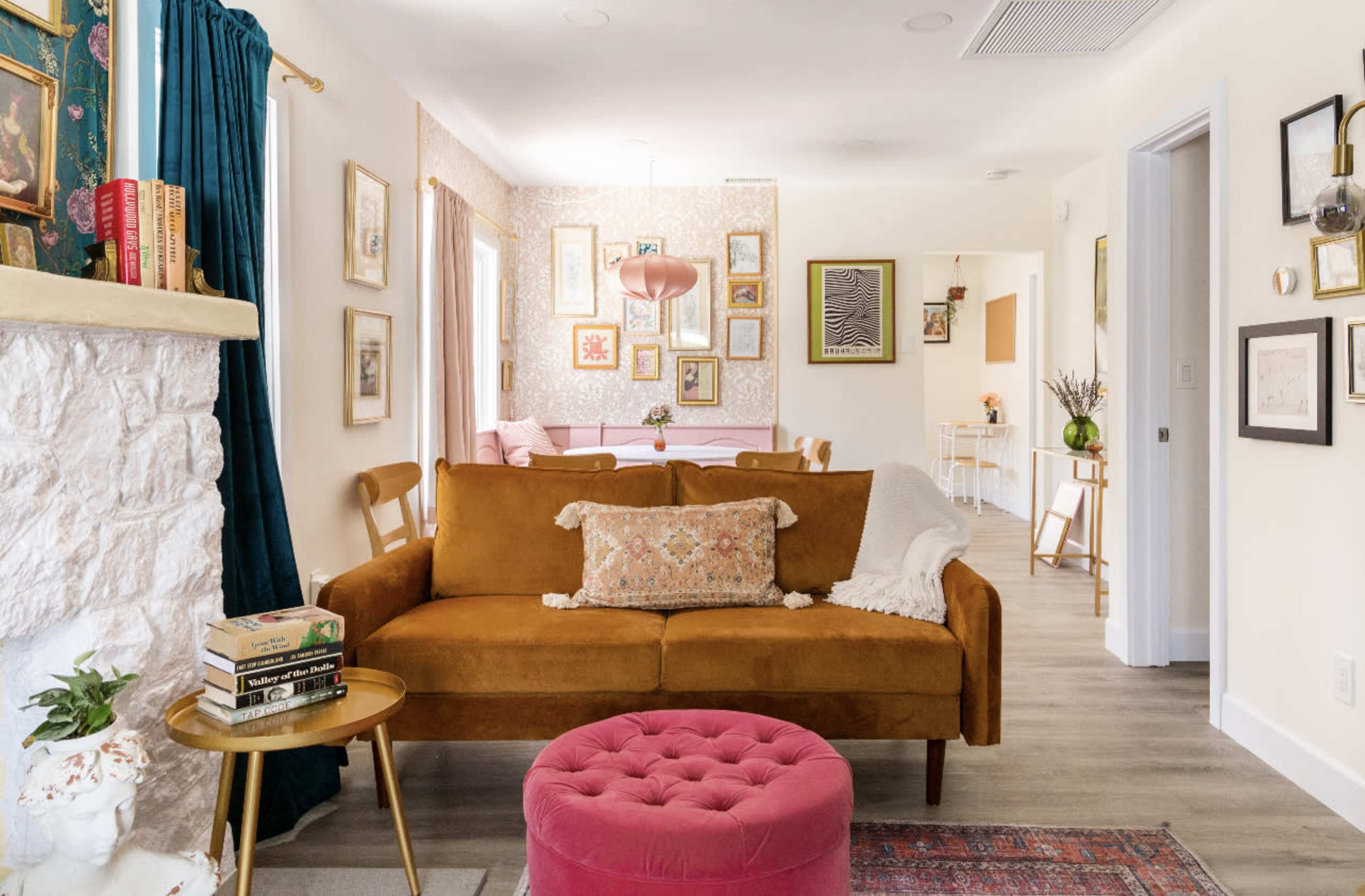 A cozy living room features a brown sofa with a decorative pillow, a round pink ottoman, and framed artwork on the walls, leading into a dining area.