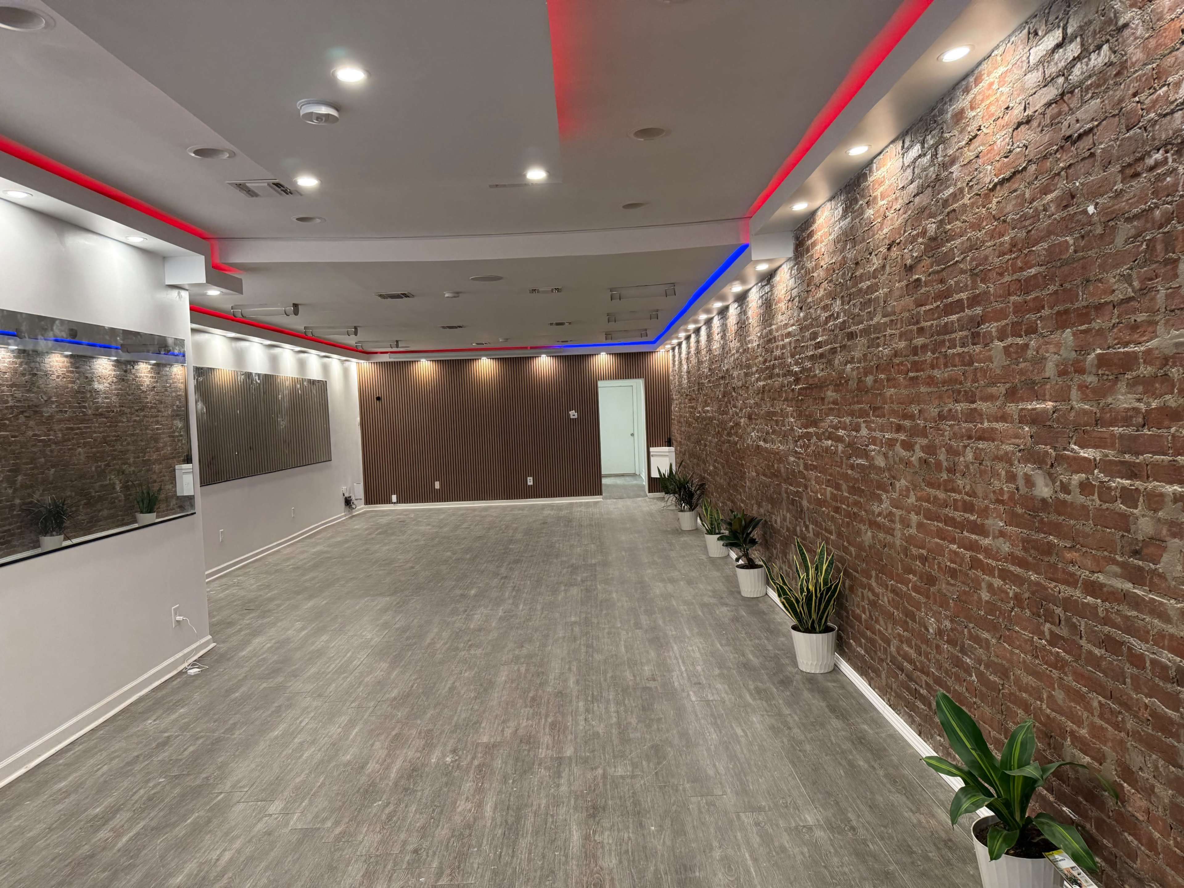The image shows an empty room with exposed brick walls, wood-paneled accents, recessed lighting, and potted plants along the floor.