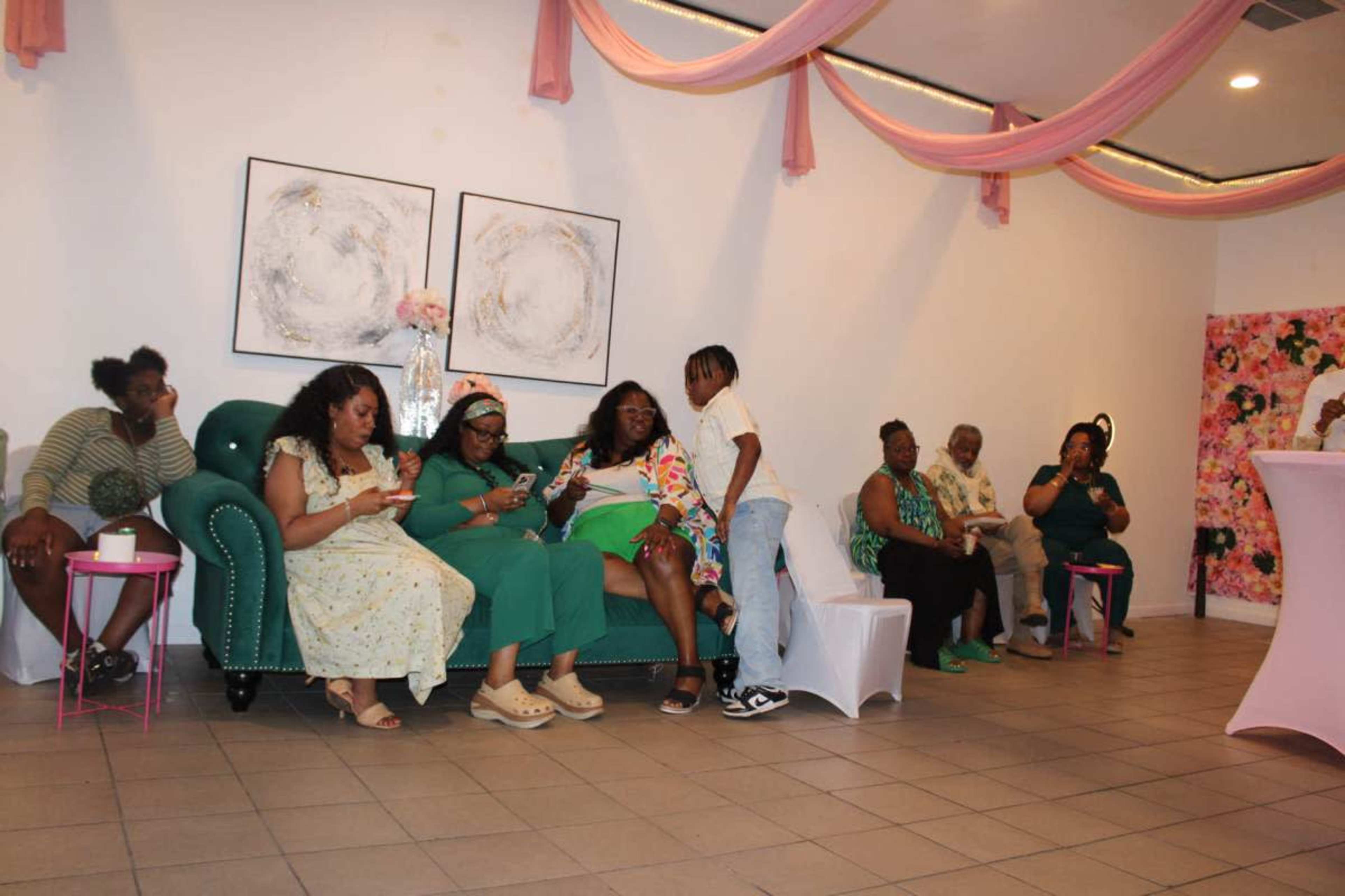 A group of people is seated and interacting in a brightly decorated room with pink drapery and floral artwork.