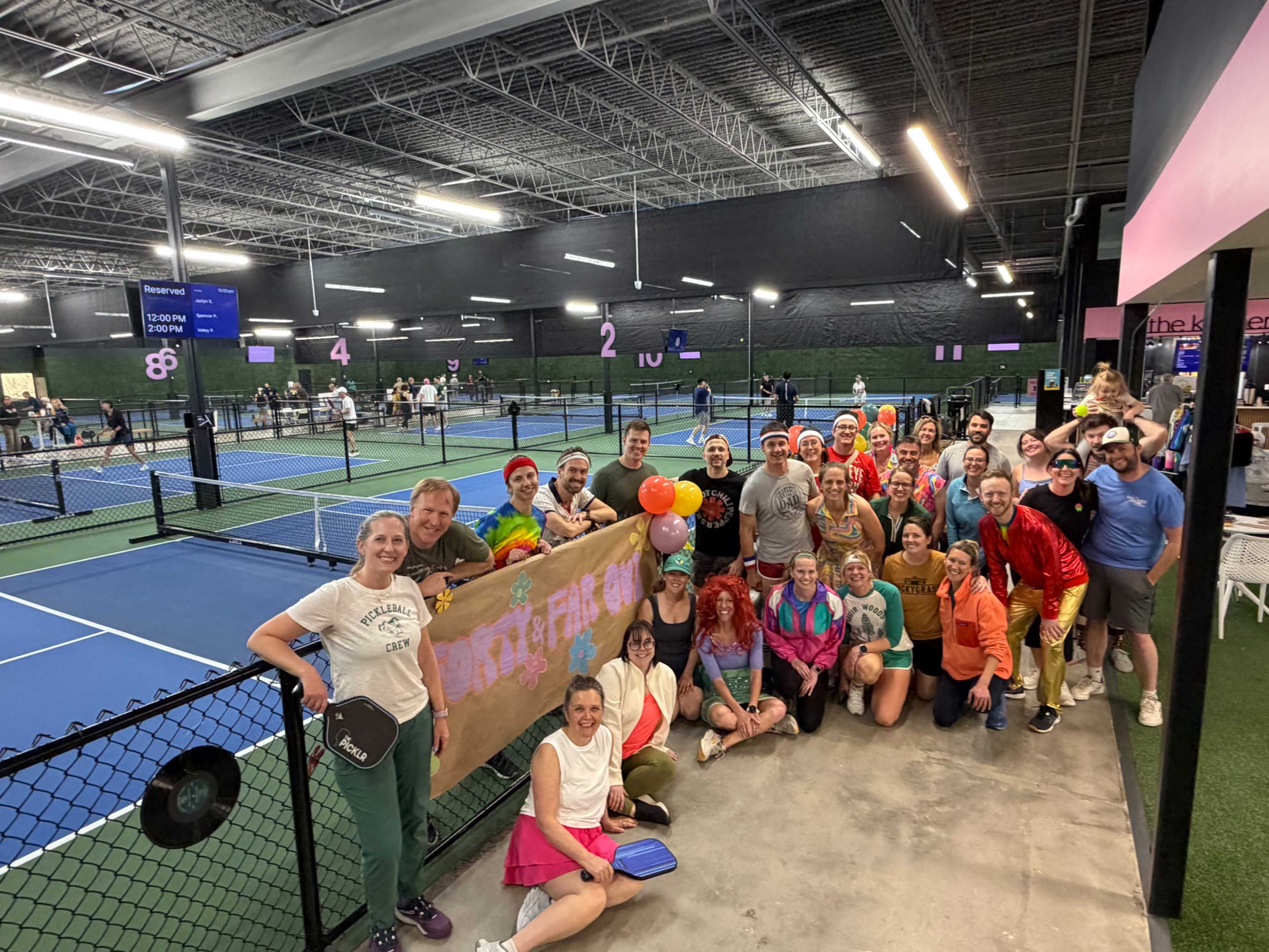A large group of people, dressed in colorful outfits, poses together in an indoor tennis facility, holding a banner with a cheerful message.