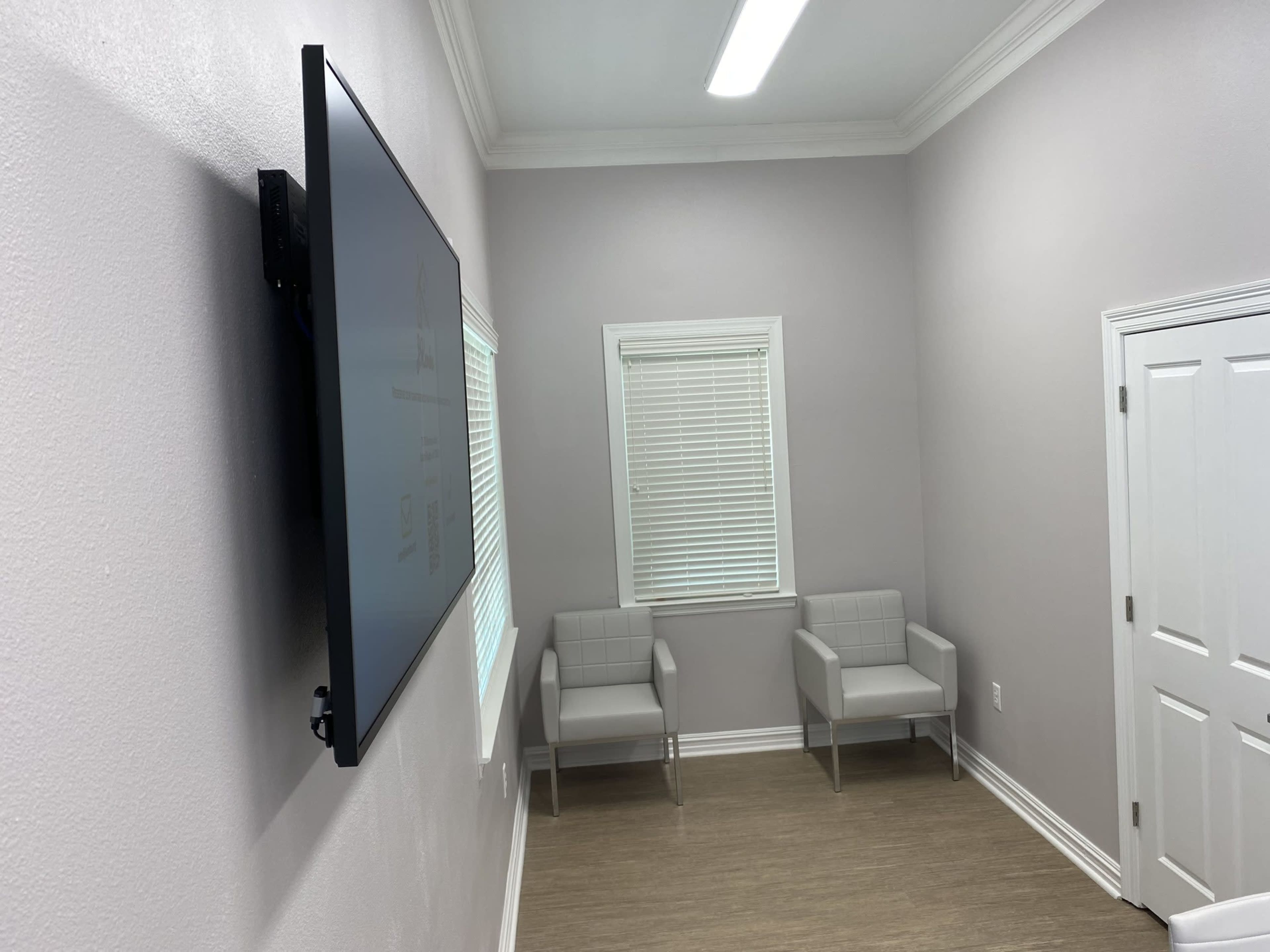 A small, empty room features two gray chairs, a wall-mounted television, and a window with blinds.