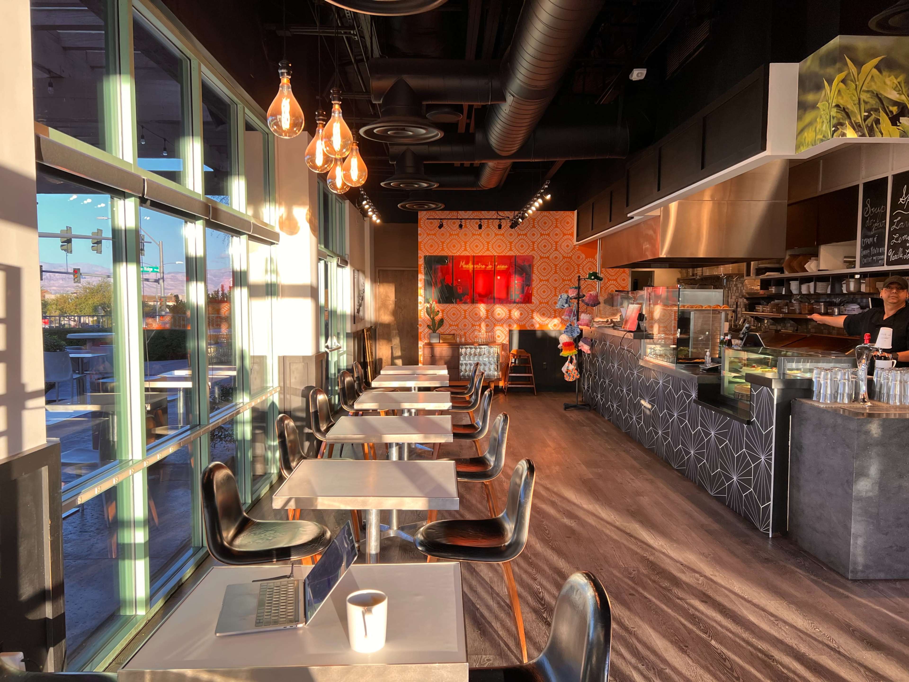A modern café with large windows letting in sunlight, featuring black chairs and tables, a coffee counter, and decorative wall patterns.