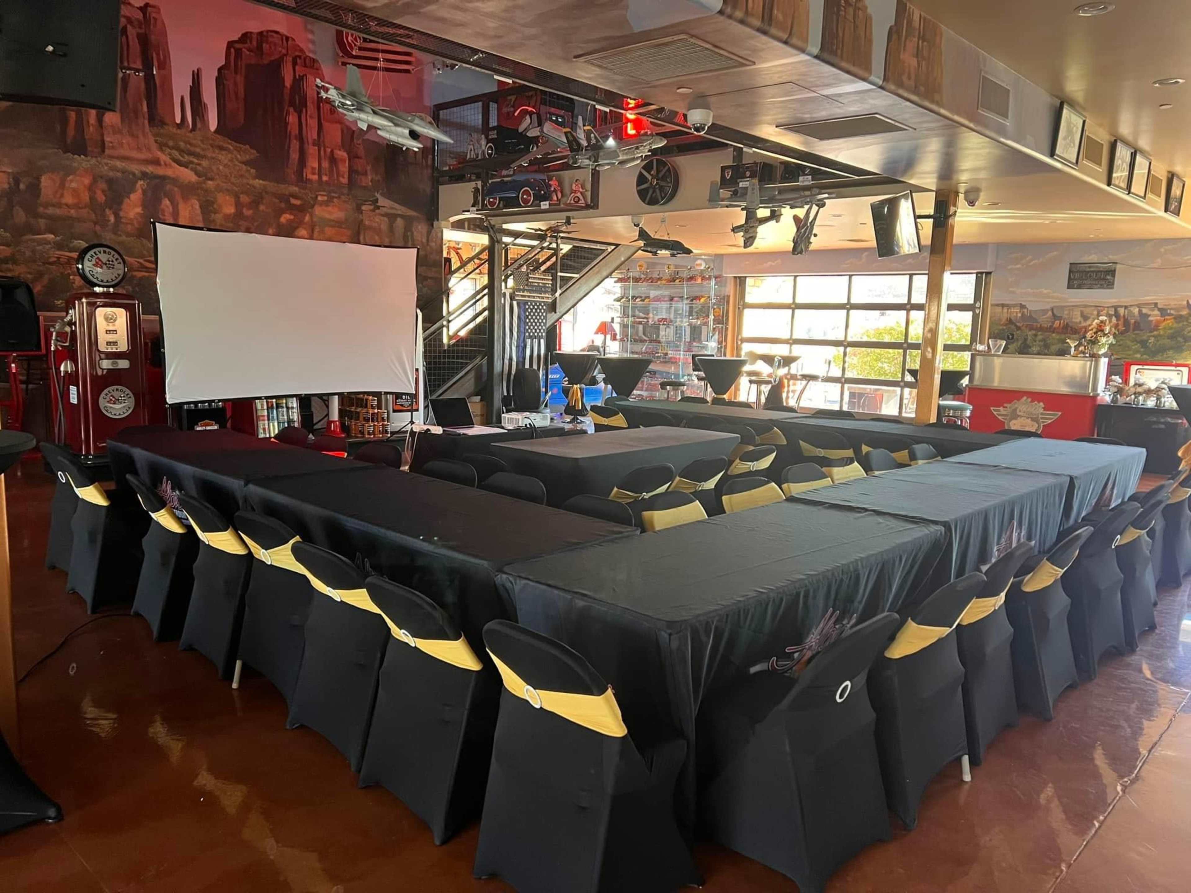 The image shows a large event space set up with rows of tables covered in black cloth, surrounded by black chairs with gold sashes, and a large screen at the front.