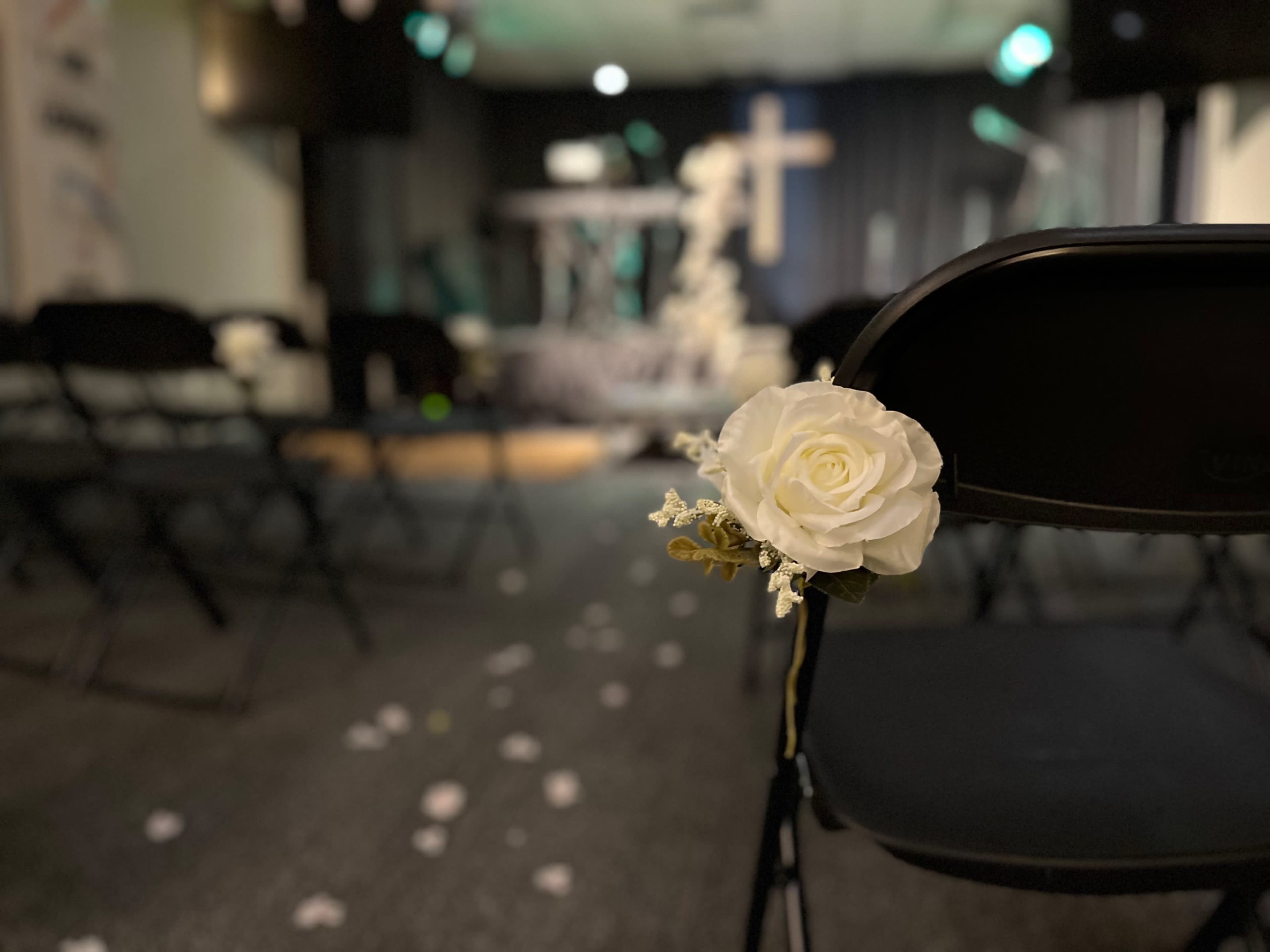 A white rose is attached to a black folding chair, with a backdrop of empty chairs and a stage in a dimly lit room.