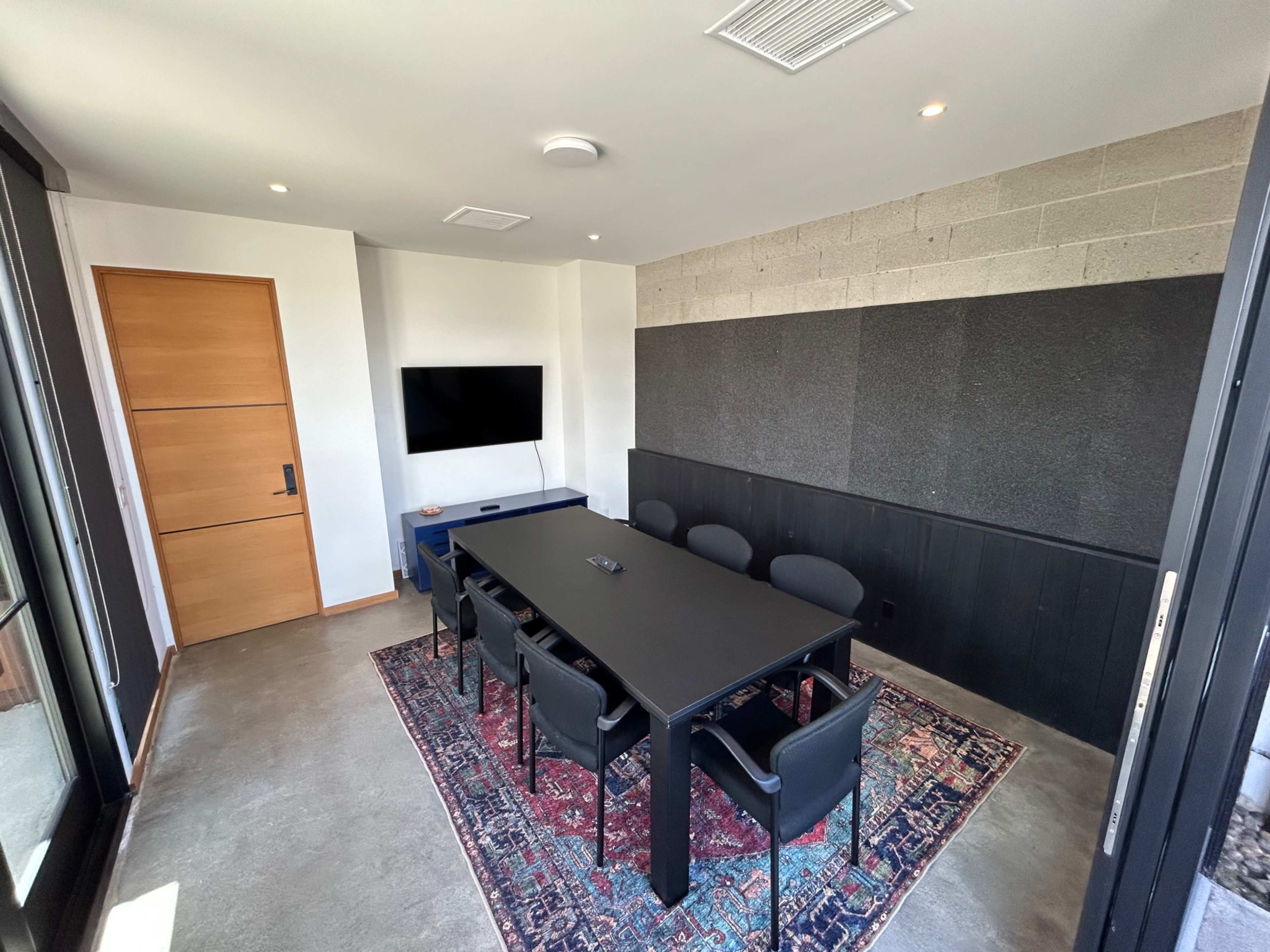 The image shows a modern conference room featuring a black table surrounded by six chairs, a wall-mounted television, and a decorative rug on the floor.