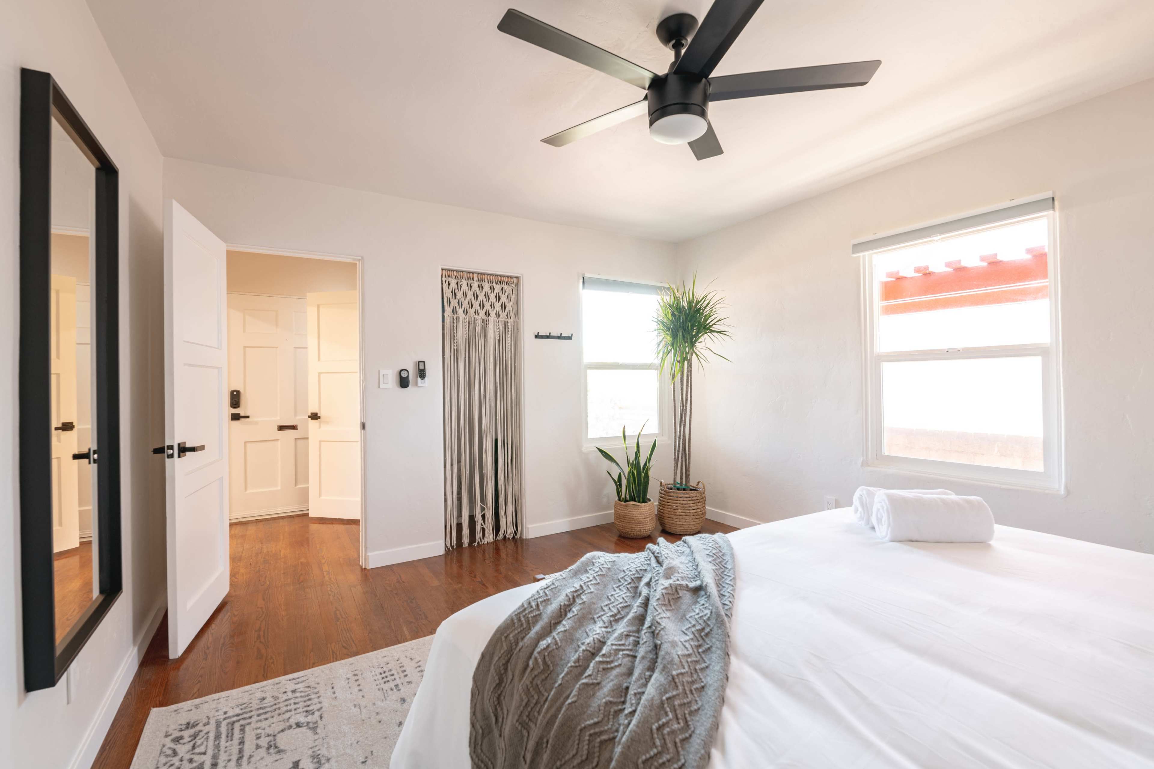 A bright bedroom features a bed with a rolled towel on it, a ceiling fan, a potted plant, and a mirror opposite the door.