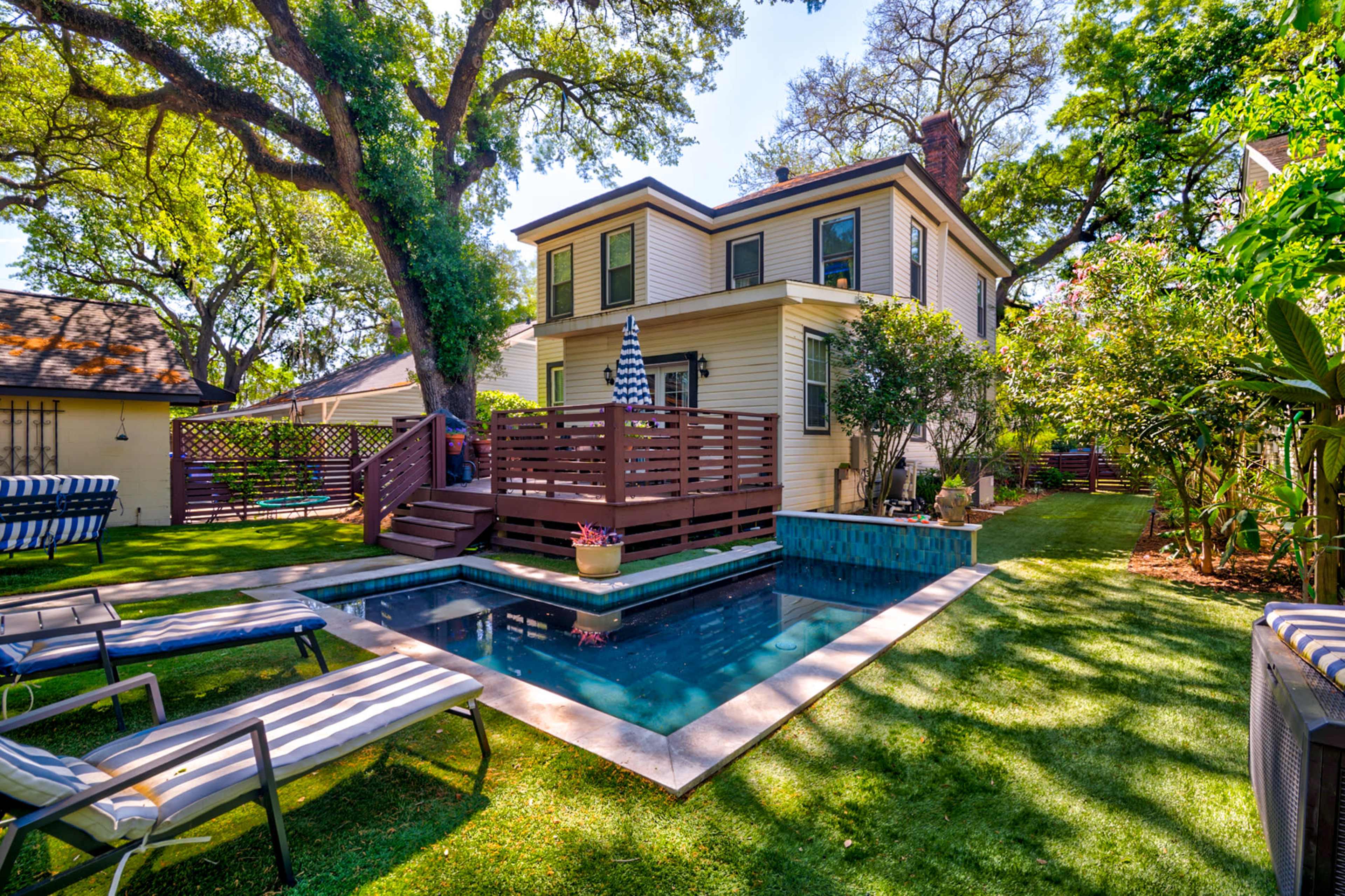 The image shows a backyard with a rectangular swimming pool surrounded by lawn chairs, a wooden deck, and a two-story house with trees and greenery nearby.