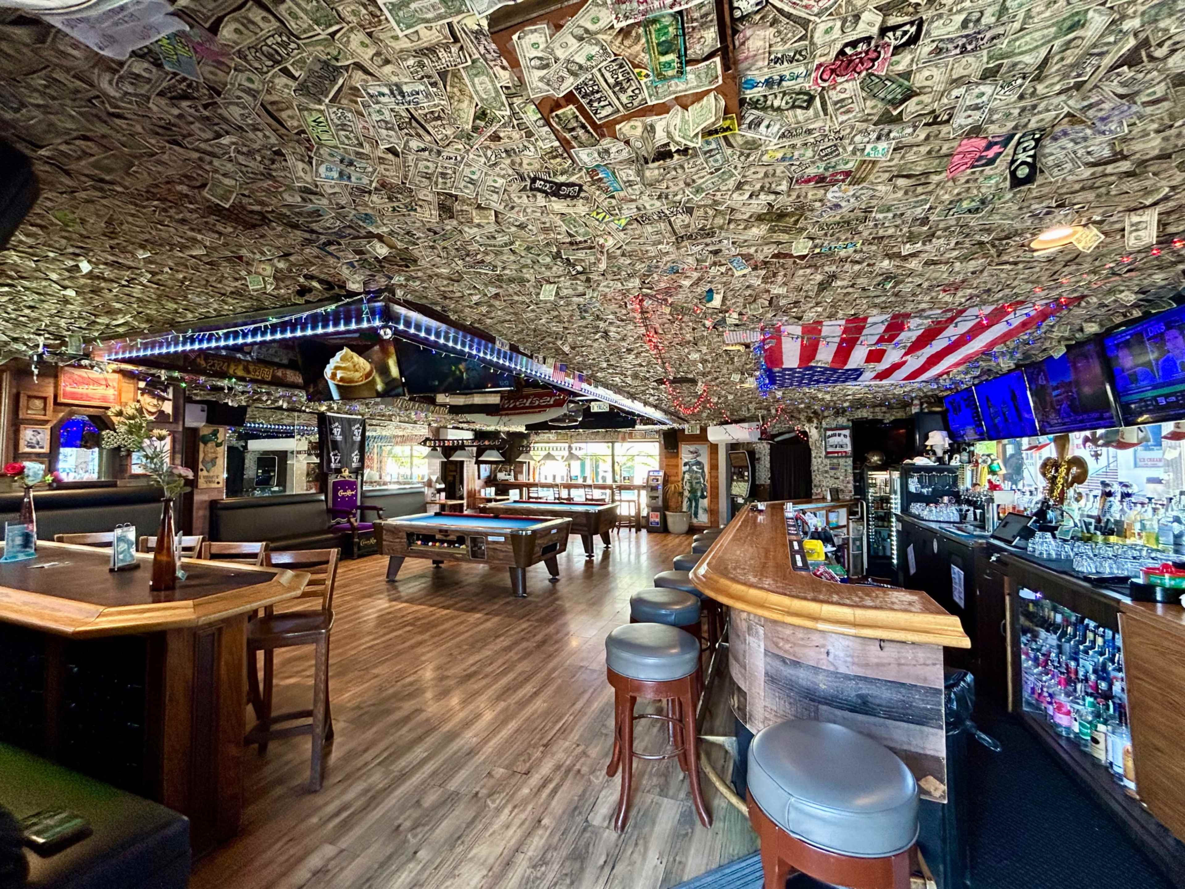 The interior of a bar features a ceiling covered in dollar bills, with pool tables, a bar area, and various decorations including string lights and an American flag.
