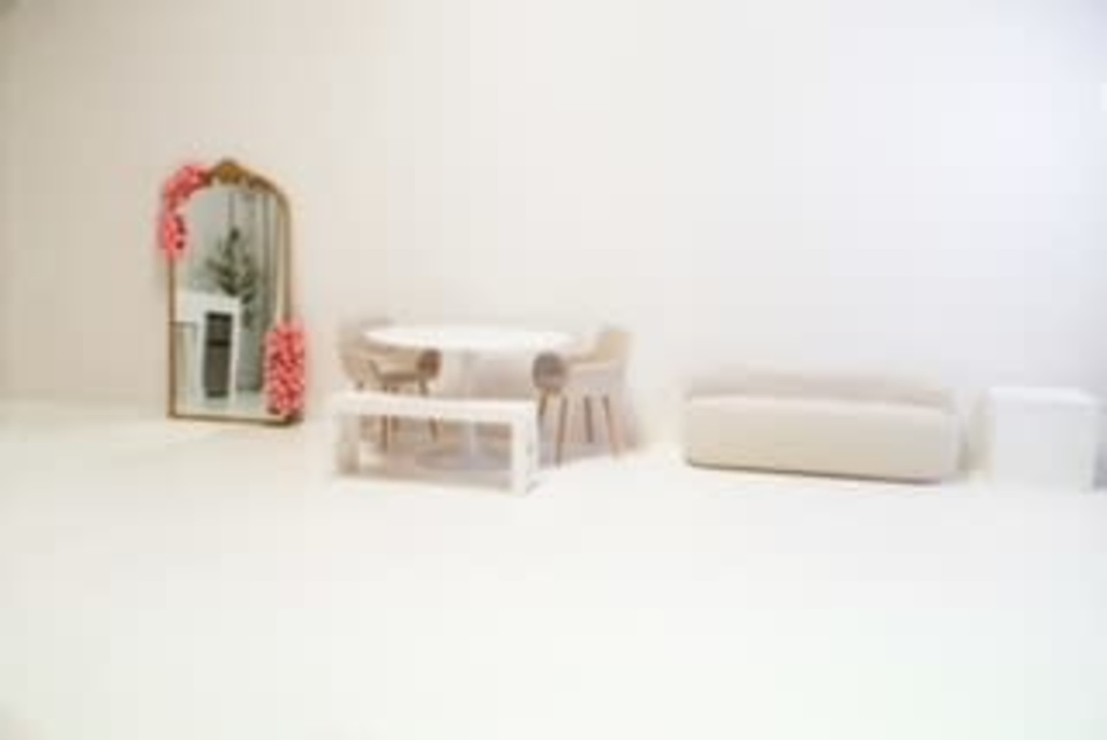 The image shows a minimalist interior space featuring a round table with chairs, a white couch, a mirror adorned with decorative elements, and a simple trash bin.