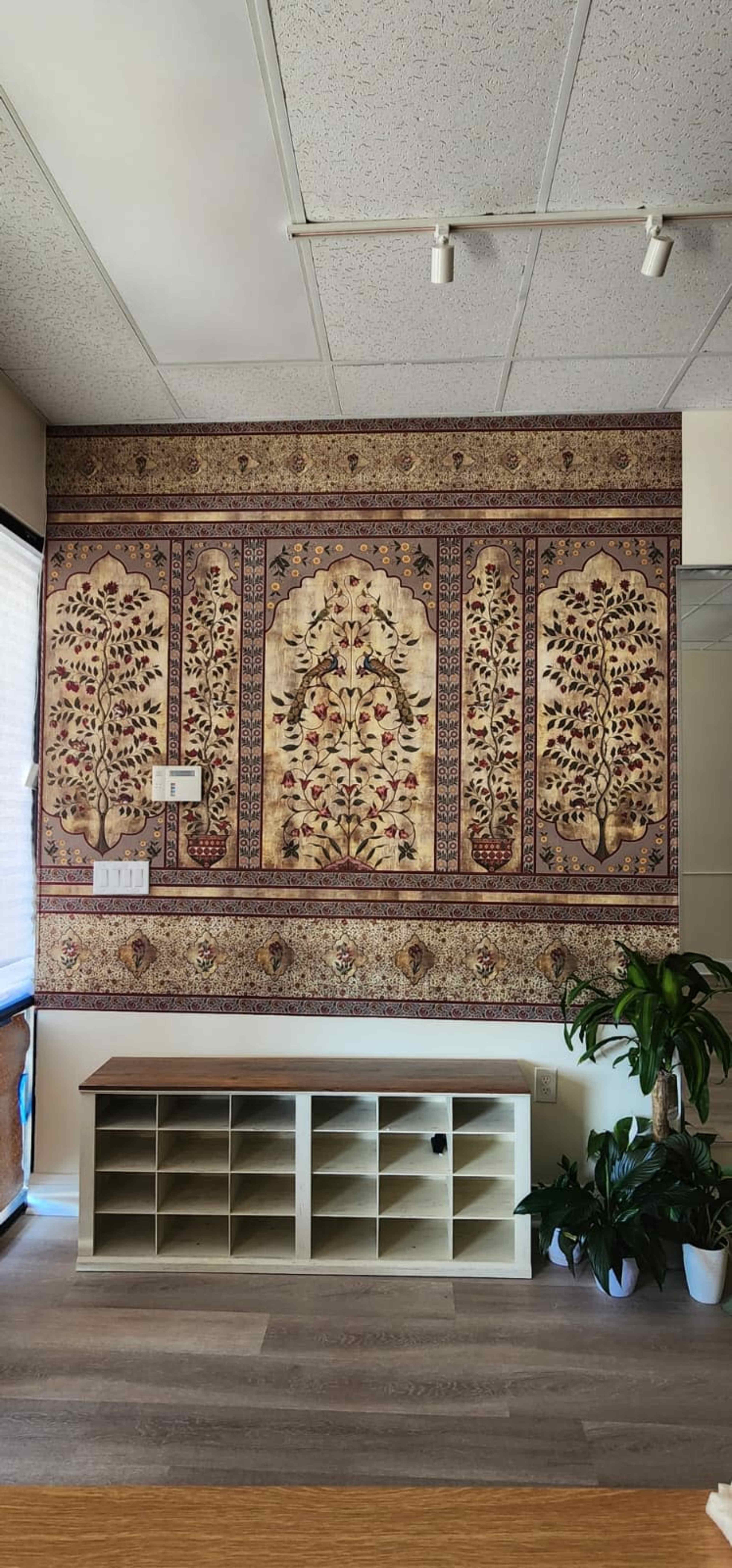 The image shows a wall adorned with a patterned textile artwork featuring floral designs, alongside a low wooden storage unit with cubbies and potted plants.