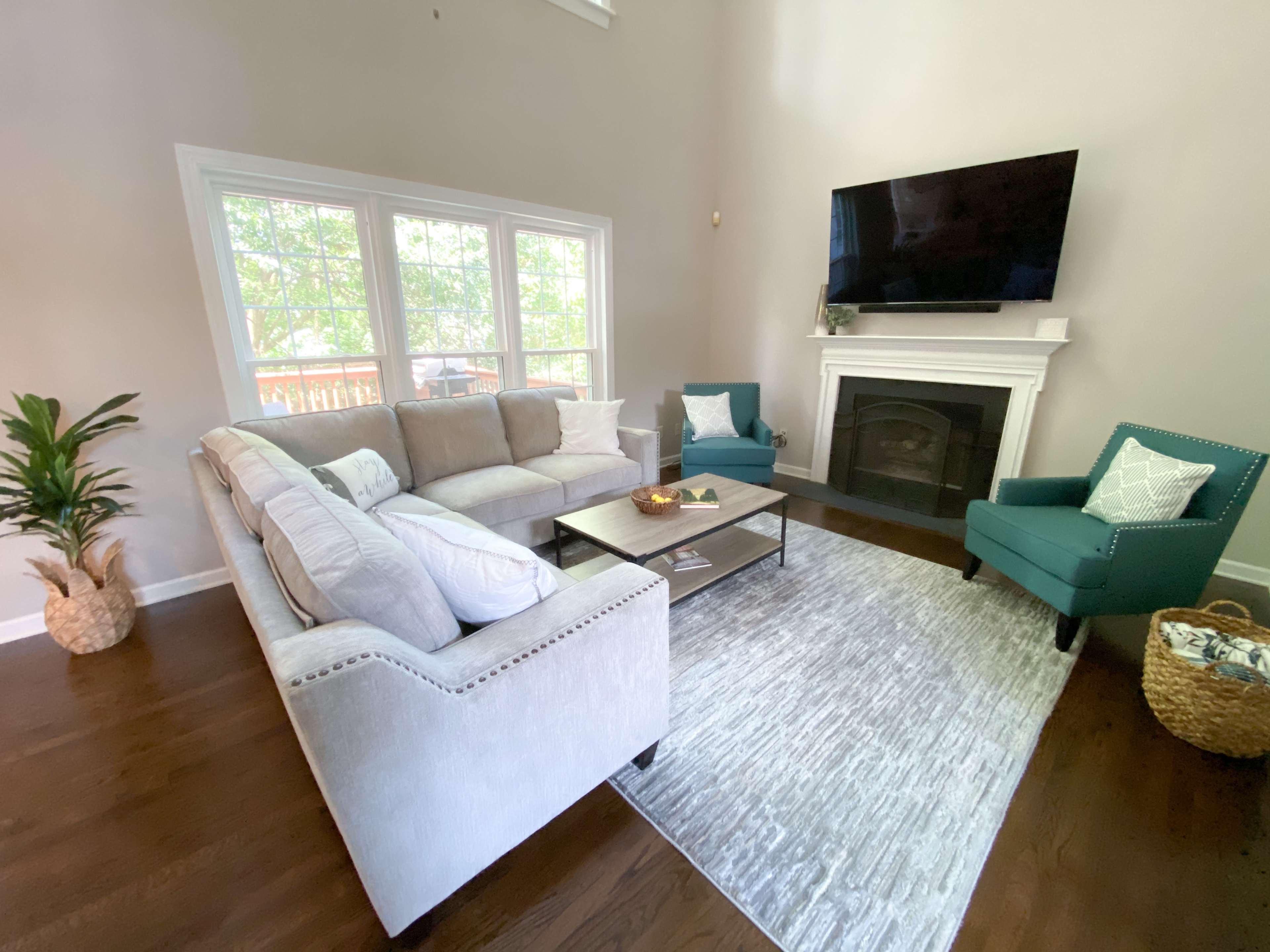 A living room features a light gray sectional sofa, teal armchairs, a coffee table, and a large television mounted above a white fireplace, complemented by natural light from three windows.