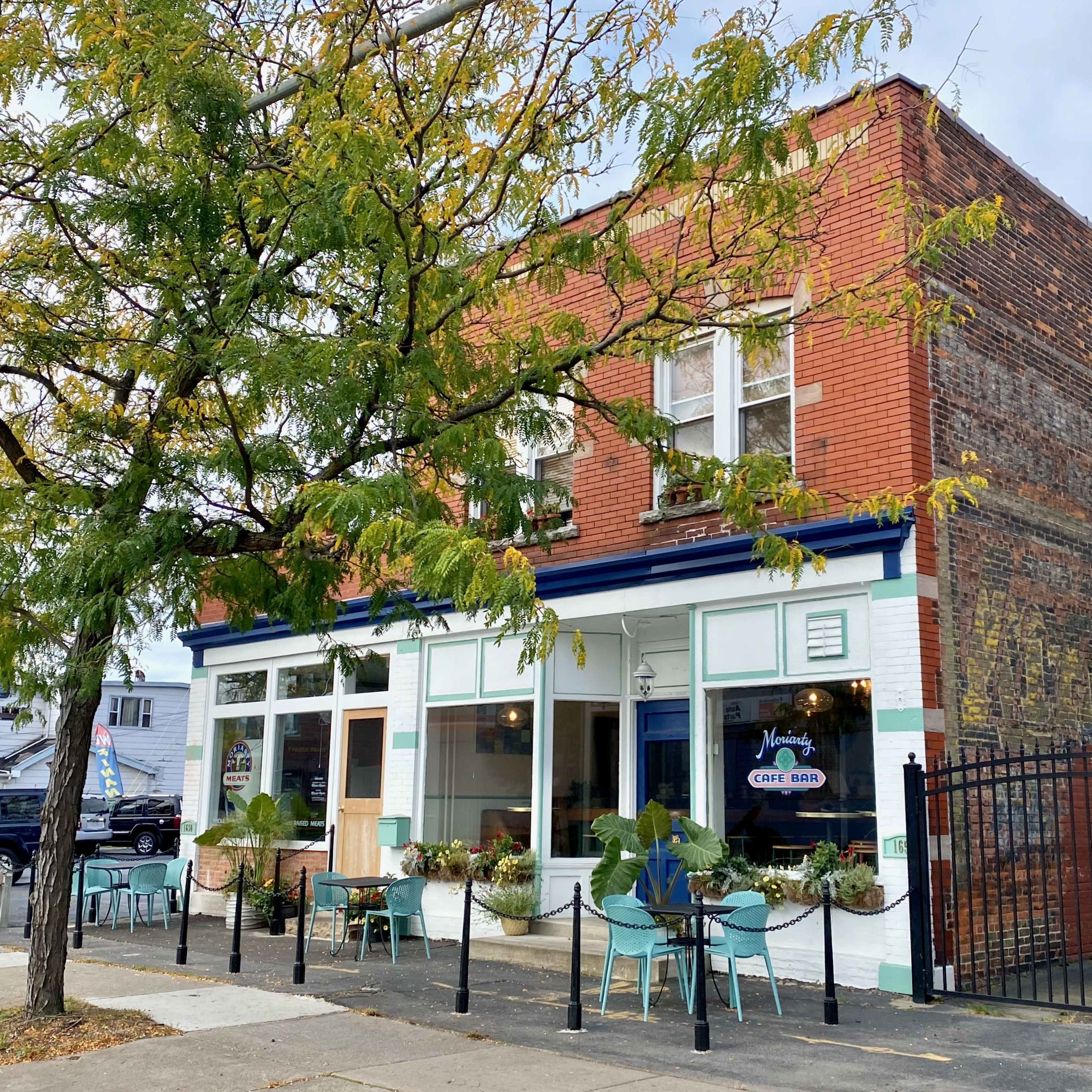 A brick building houses a café with large windows and outdoor seating, framed by a tree with yellow leaves.