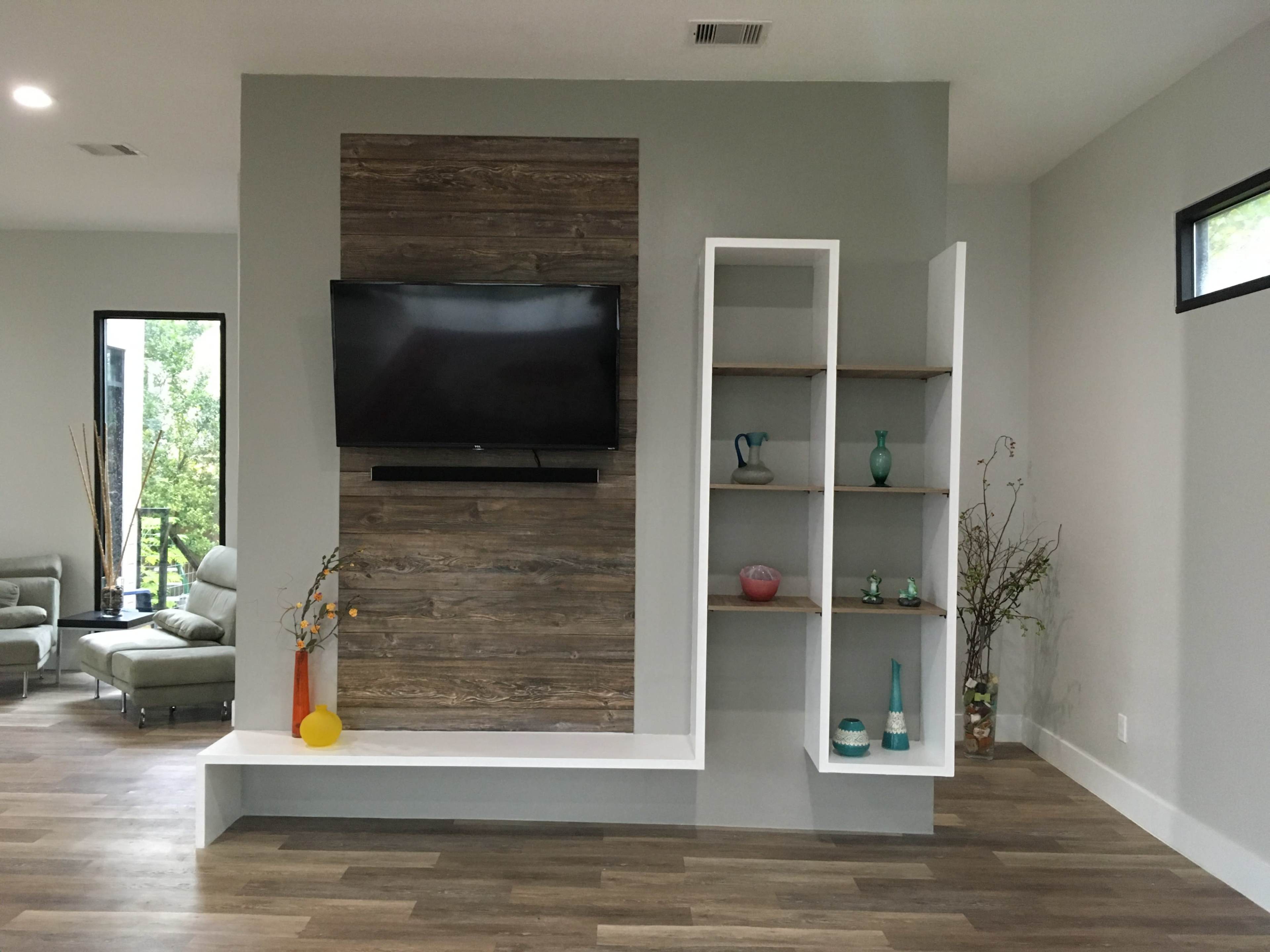 The image shows a modern living room with a wall-mounted television, a wooden accent wall, and a shelving unit displaying decorative items.
