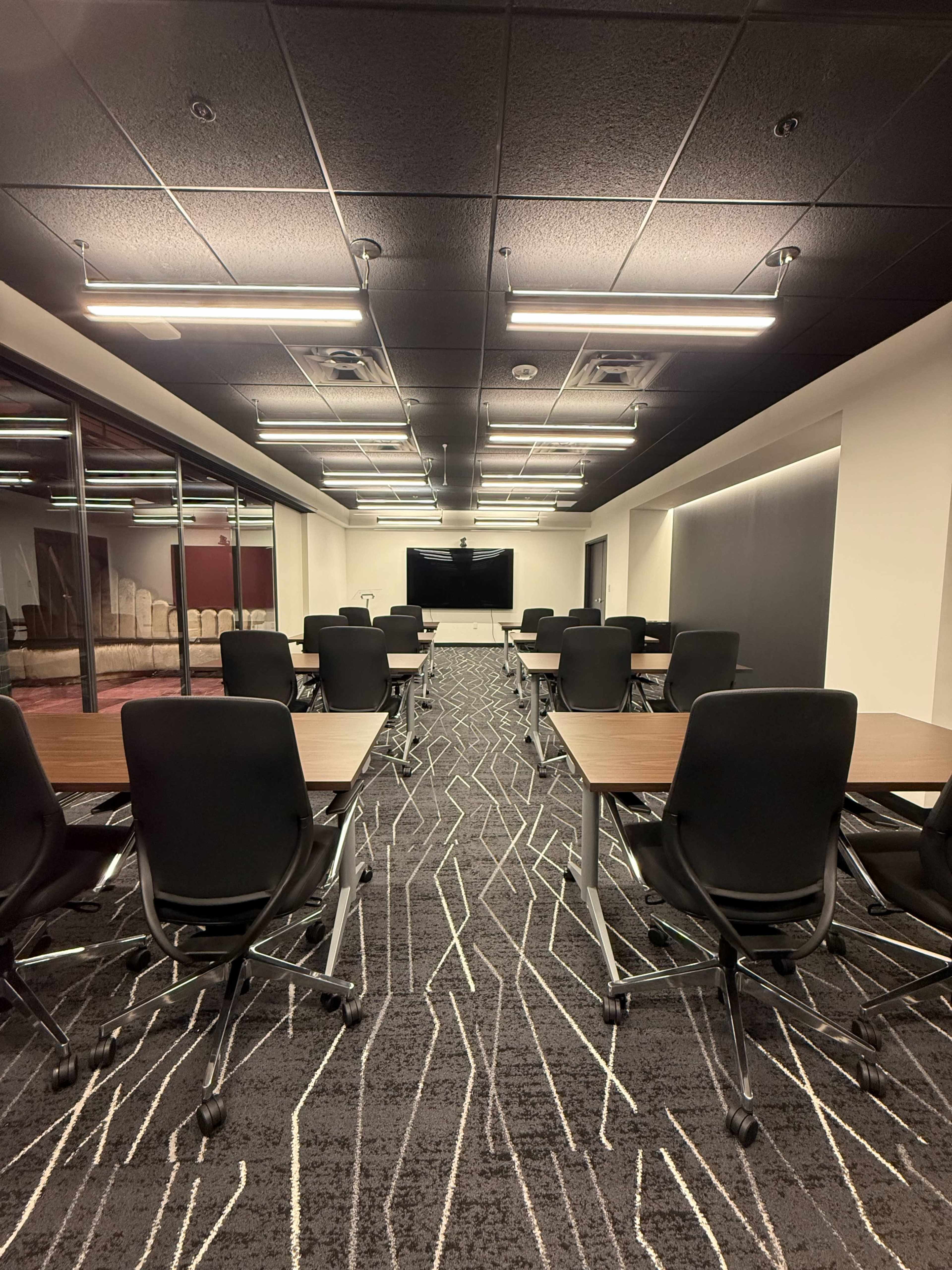 The image shows a modern conference room with rows of tables and chairs facing a large wall-mounted screen.