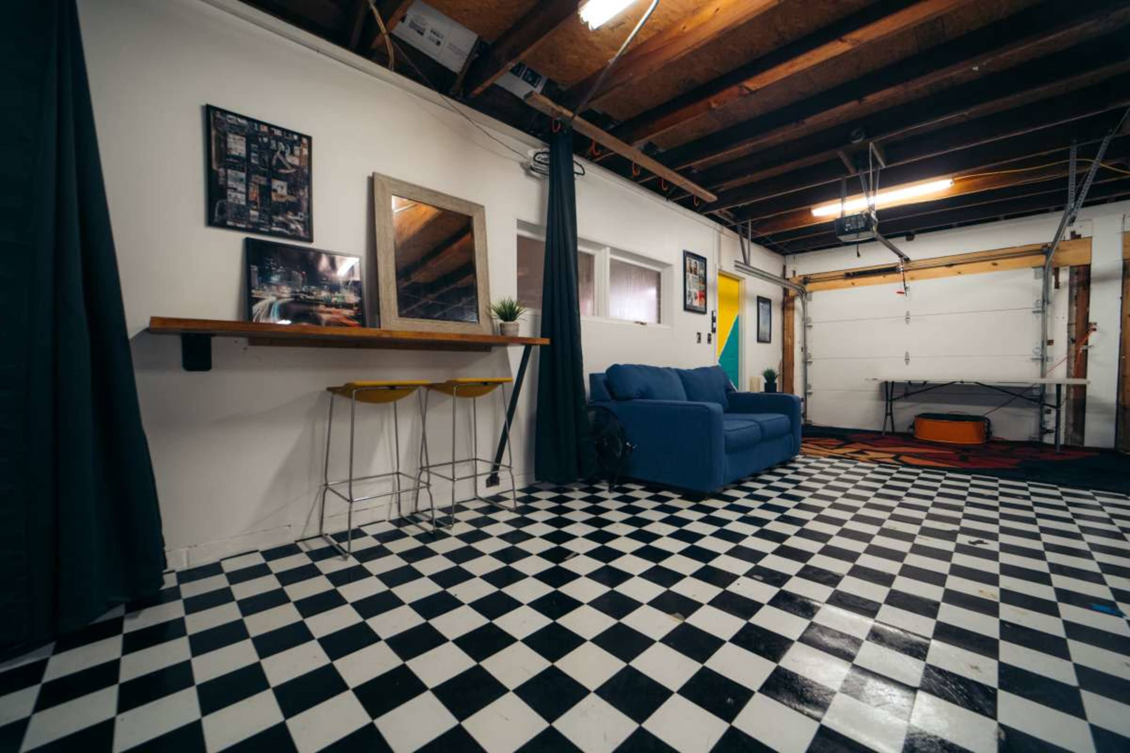 A garage converted into a small living space, featuring a blue couch, a bar-style table with two stools, and a checkerboard floor in black and white.