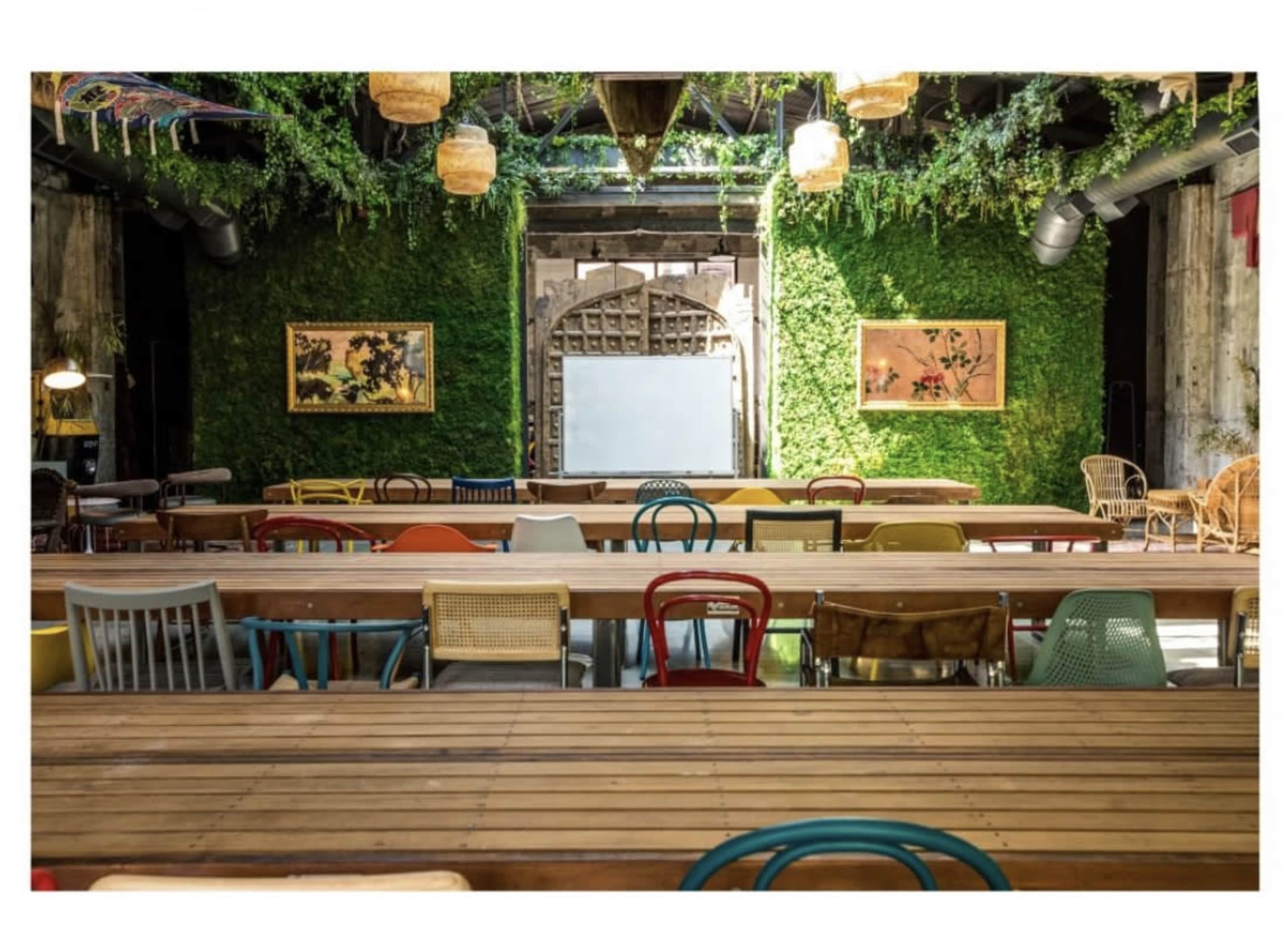 An indoor space with long wooden tables arranged for seating, surrounded by greenery-covered walls and decorative artwork.