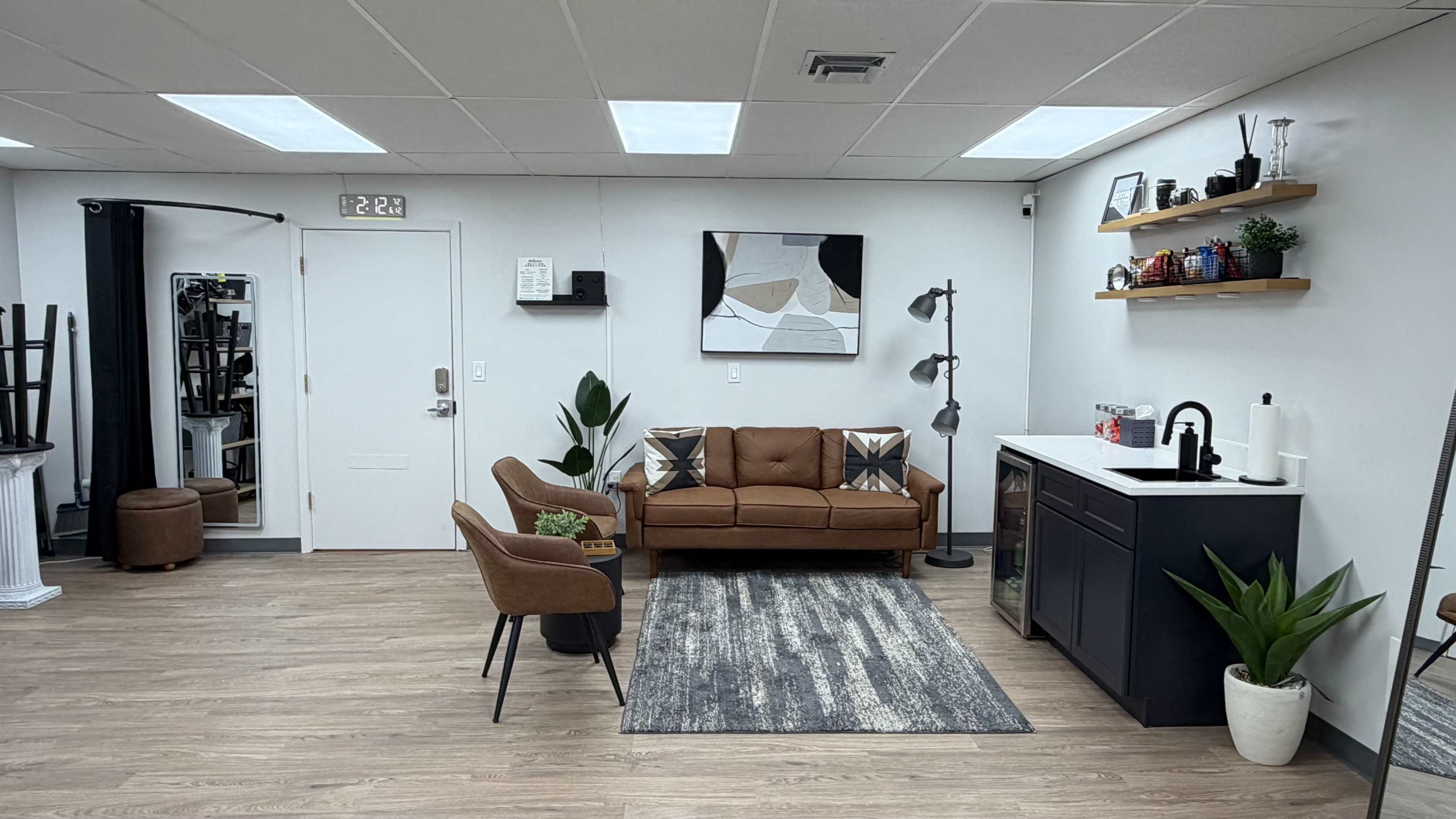 The image shows a modern interior space featuring a brown sofa, two chairs, a small kitchen area, and decorative shelves on the walls.
