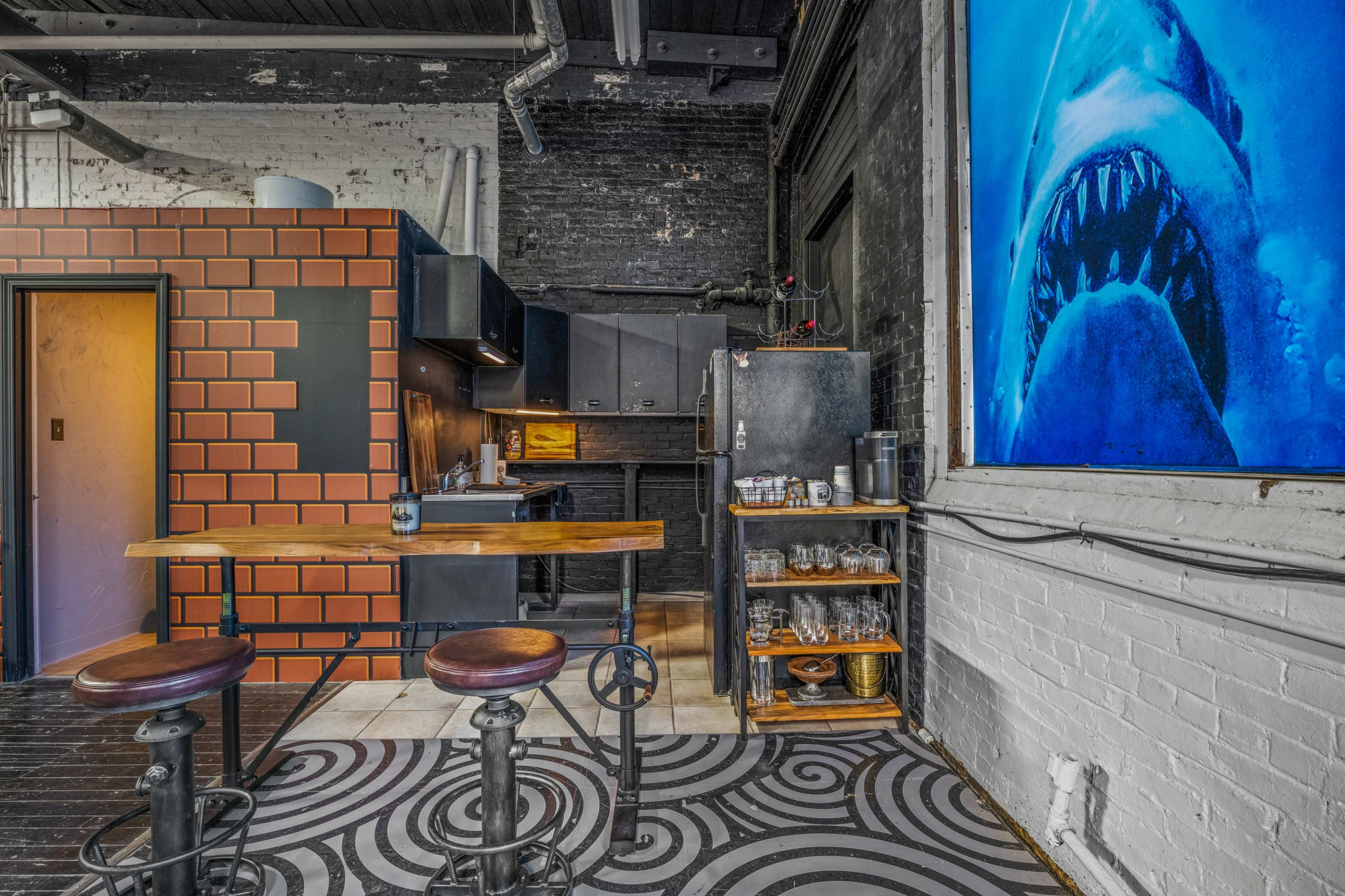 A modern kitchen with dark cabinetry, a distinctive brick-patterned wall, a wooden table with industrial-style stools, and a large wall-mounted screen displaying a shark.