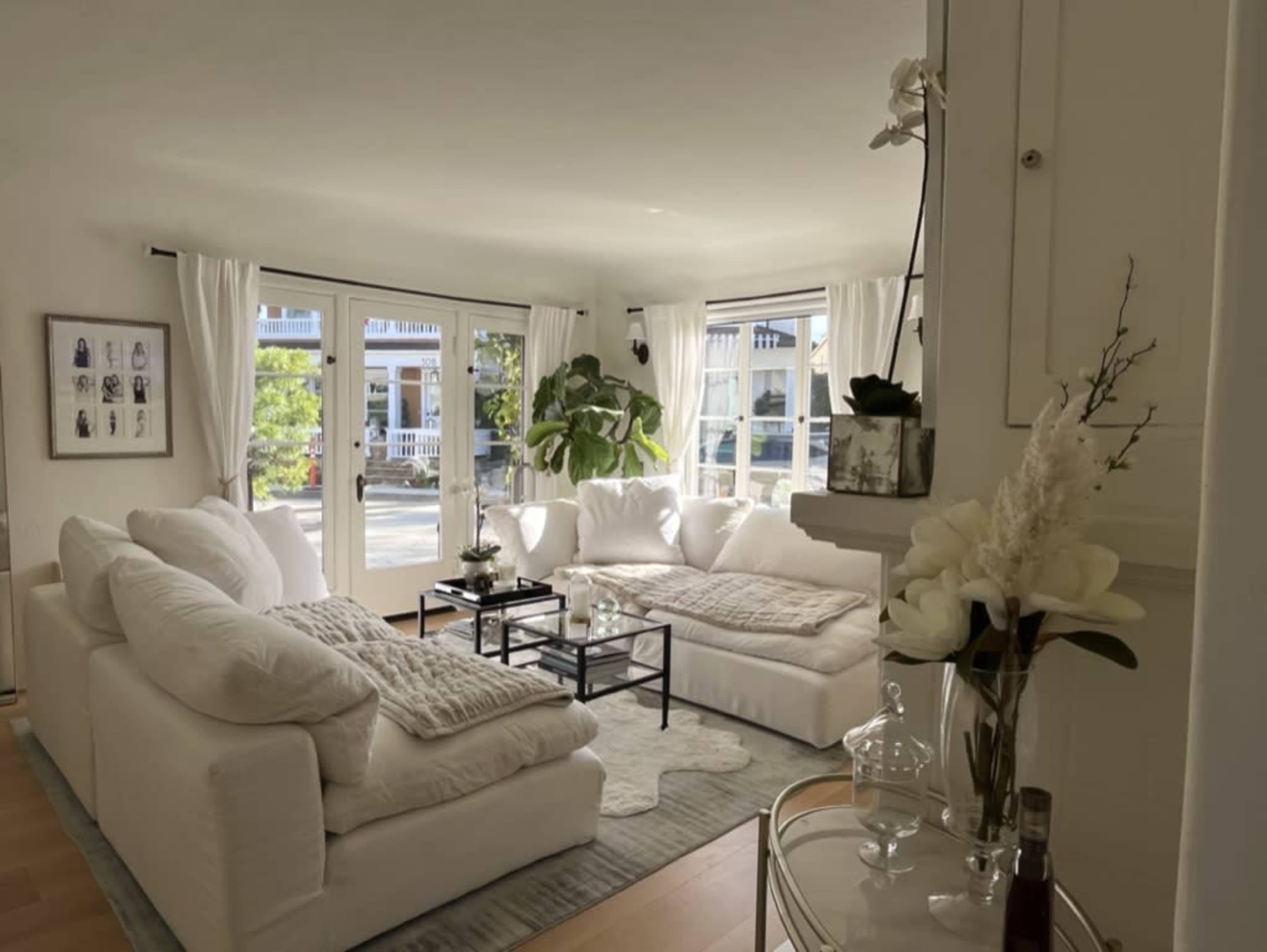 A bright living room features a large white sectional sofa, a coffee table with decorative items, and large windows allowing natural light to enter.