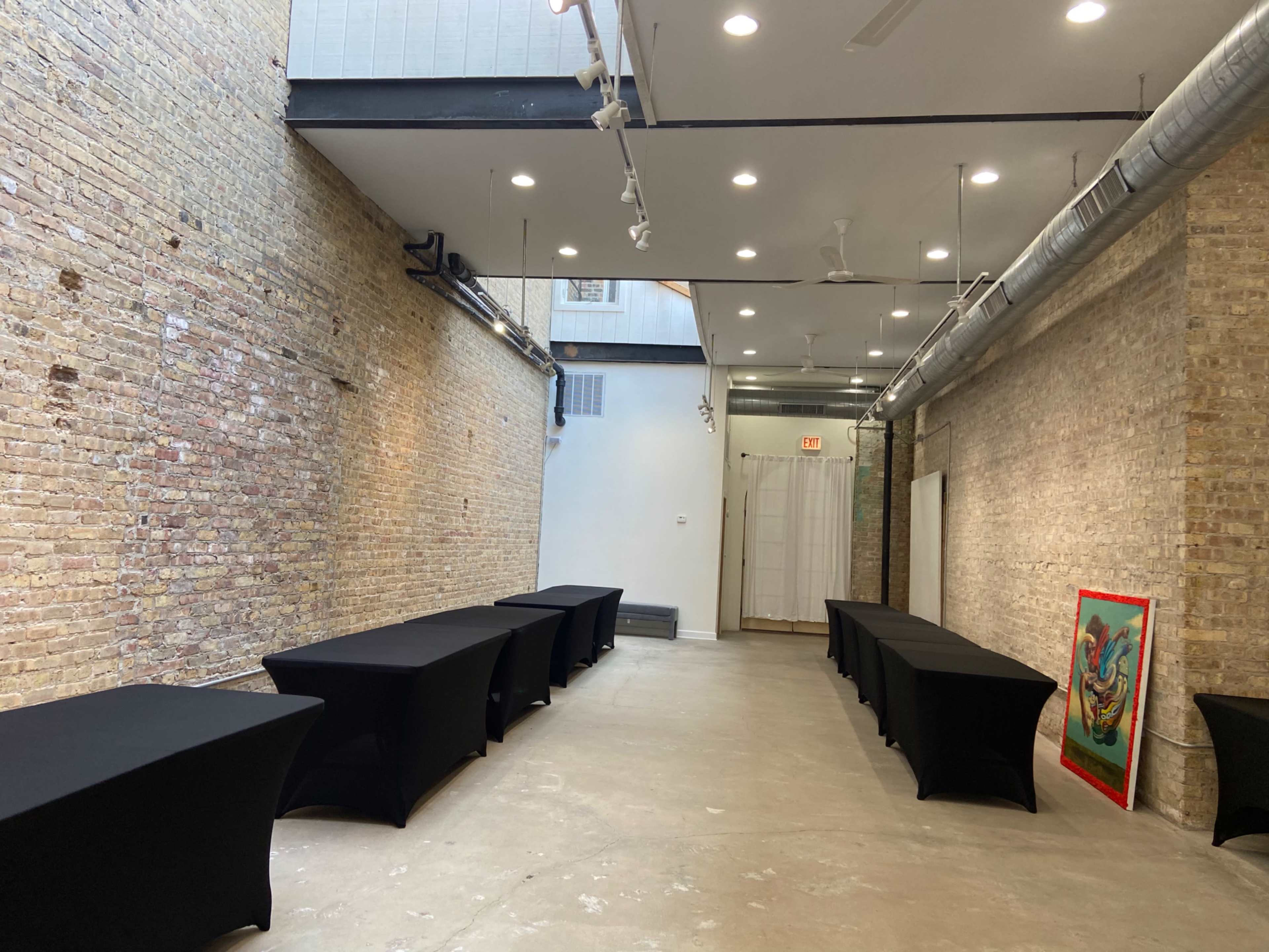 The scene shows a spacious room with exposed brick walls, several black-covered tables arranged along the sides, and a colorful artwork framed and displayed at one end.