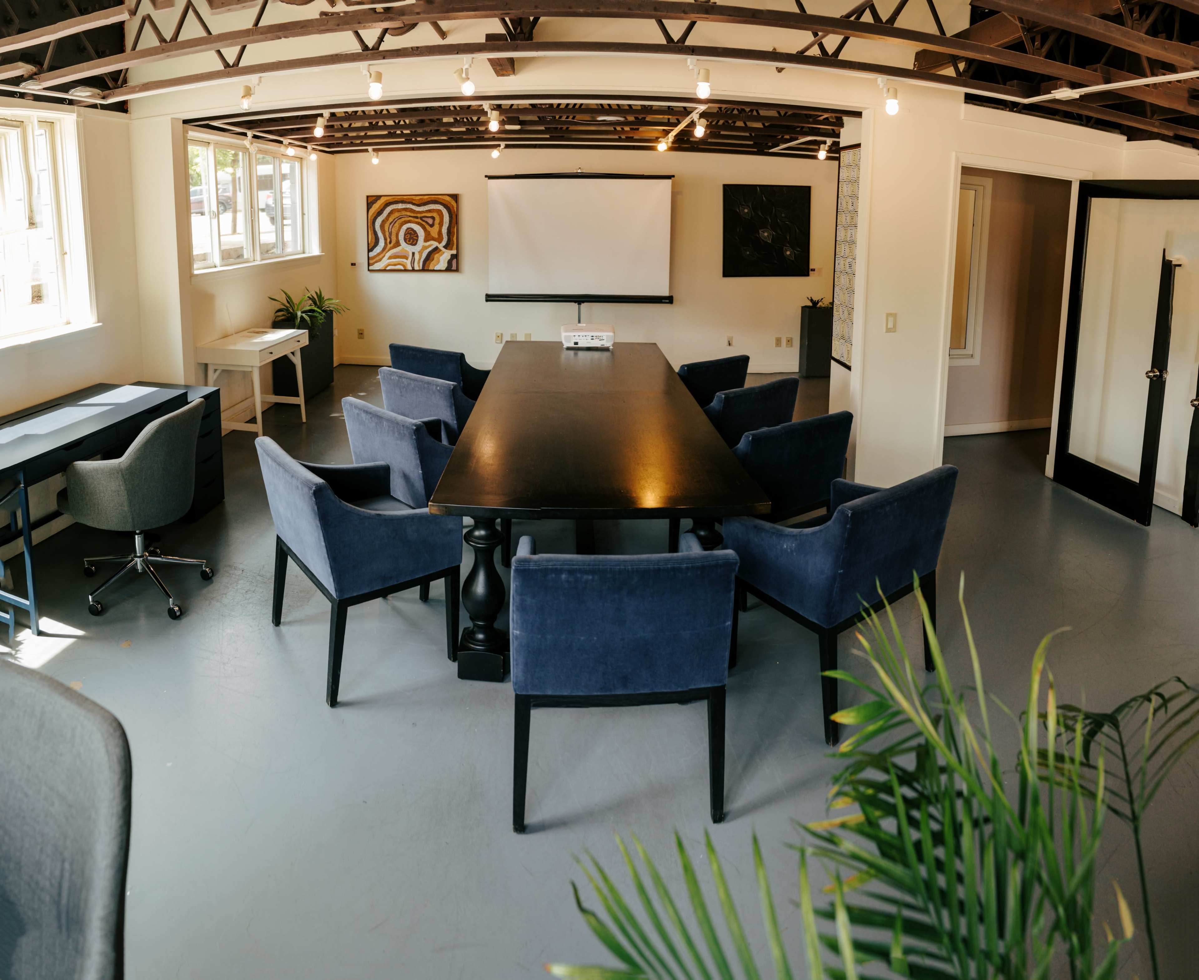 The image shows a modern conference room with a large table surrounded by upholstered chairs, white walls, and artwork on the walls.
