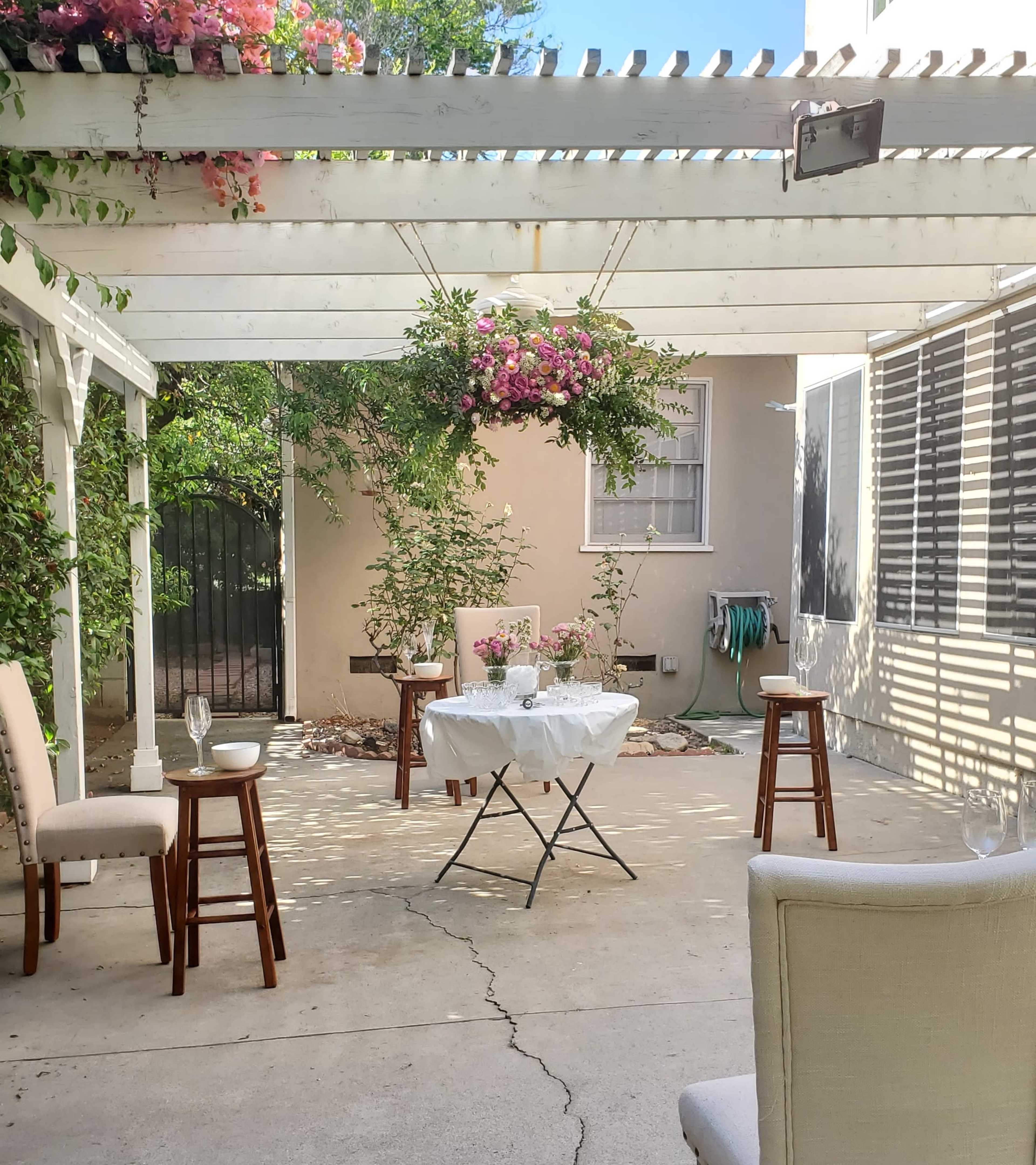 A small patio features a white table set with flowers and surrounded by four chairs and two stools under a pergola with hanging plants.