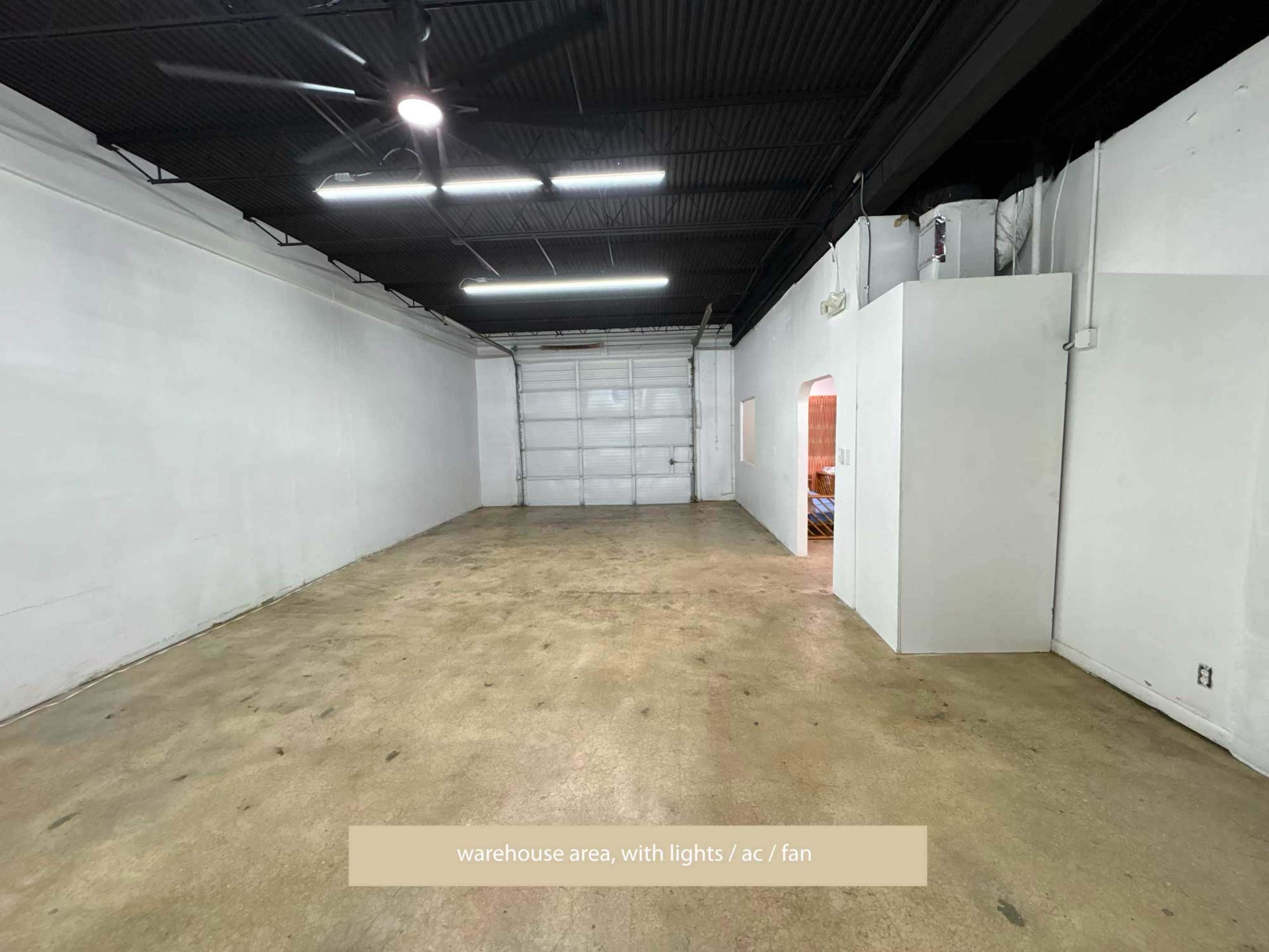The image shows a spacious warehouse area featuring a concrete floor, overhead lights, and ventilation from a fan.