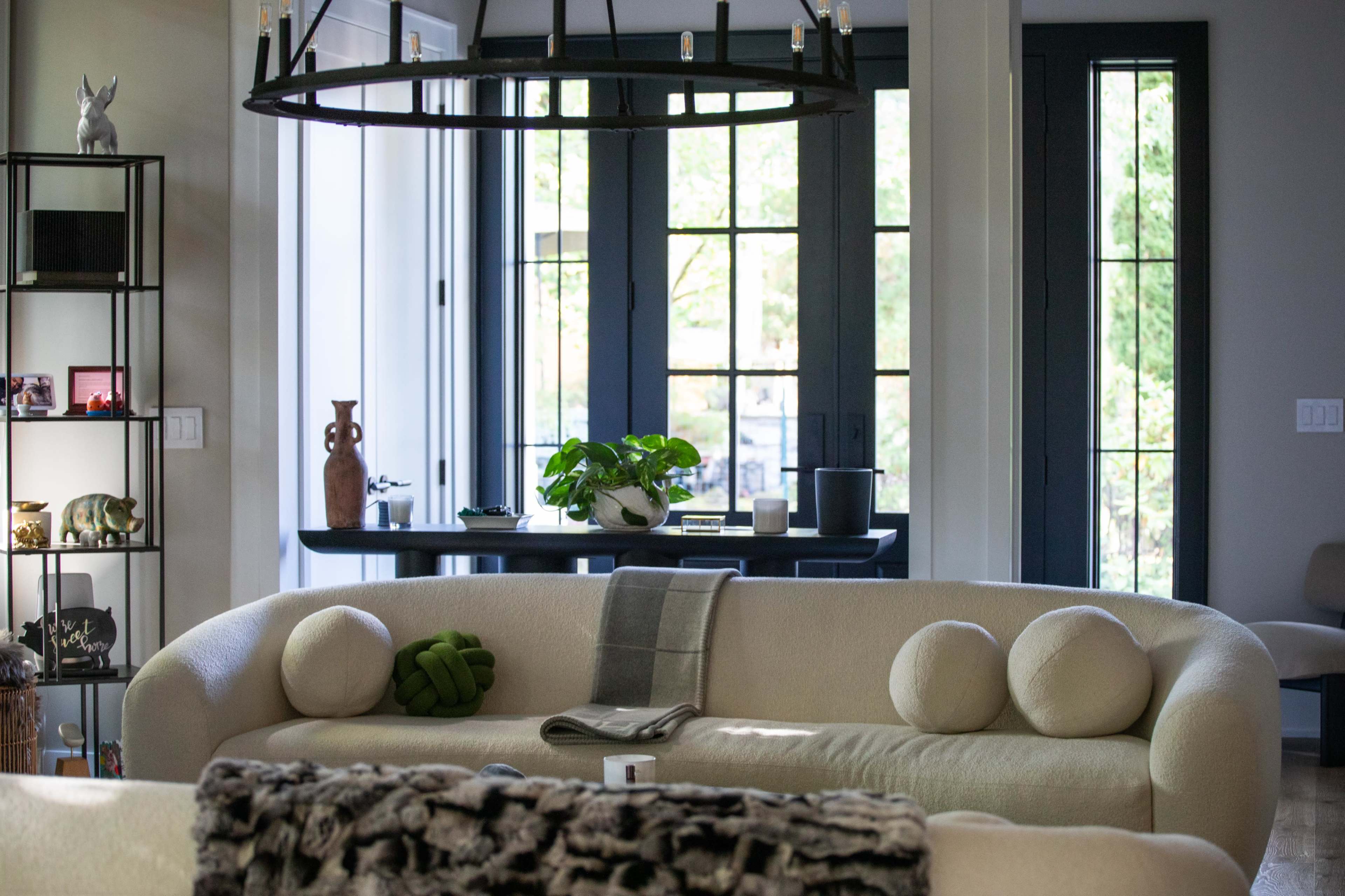 The image shows a modern living room with a rounded beige sofa, a coffee table, and large windows allowing natural light to enter.