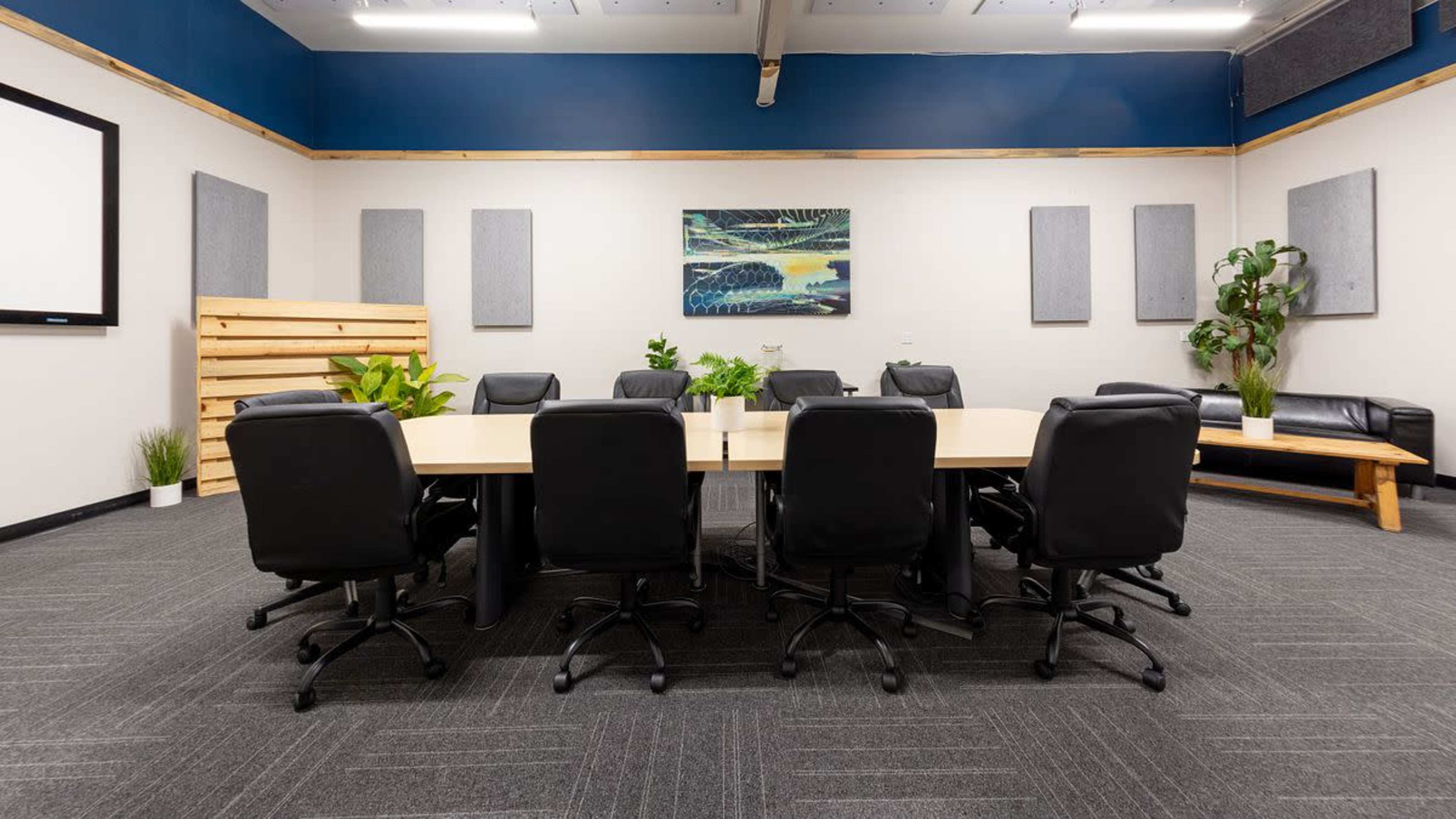 A conference room features a large table surrounded by black chairs, with potted plants and artwork on the walls.