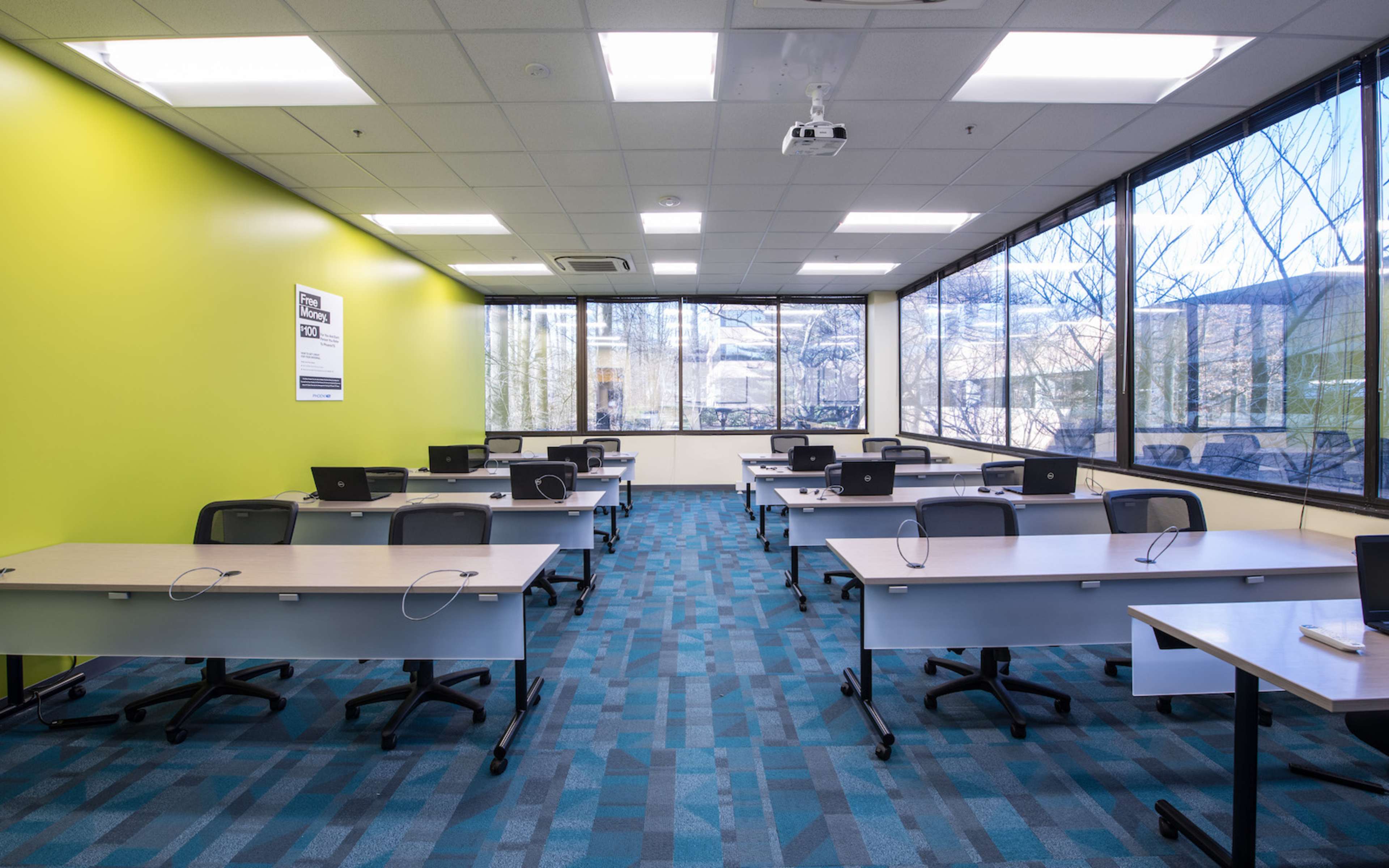 The image shows a modern classroom with several tables and chairs arranged in a grid, equipped with laptops, and large windows letting in natural light.
