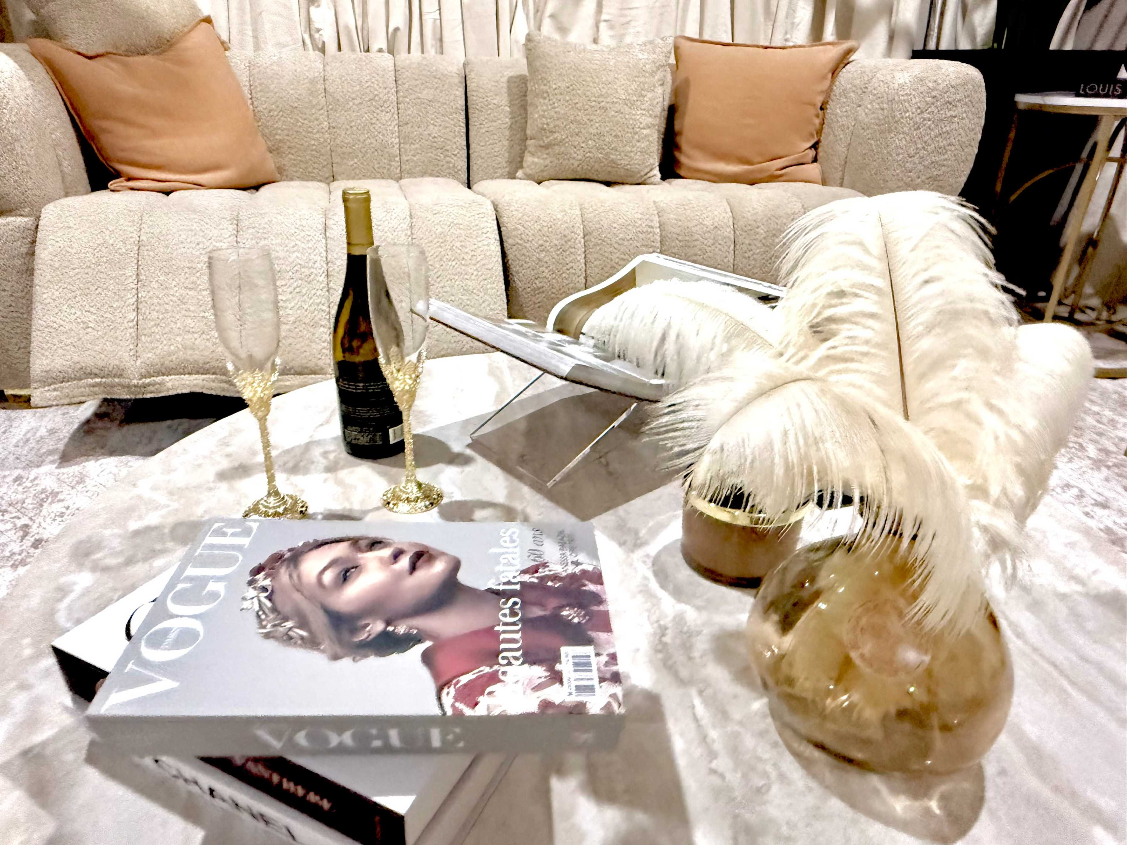 The image features a plush beige sofa with orange cushions, a coffee table adorned with a Vogue magazine, a bottle of champagne, two flute glasses, and decorative feathers in a vase.