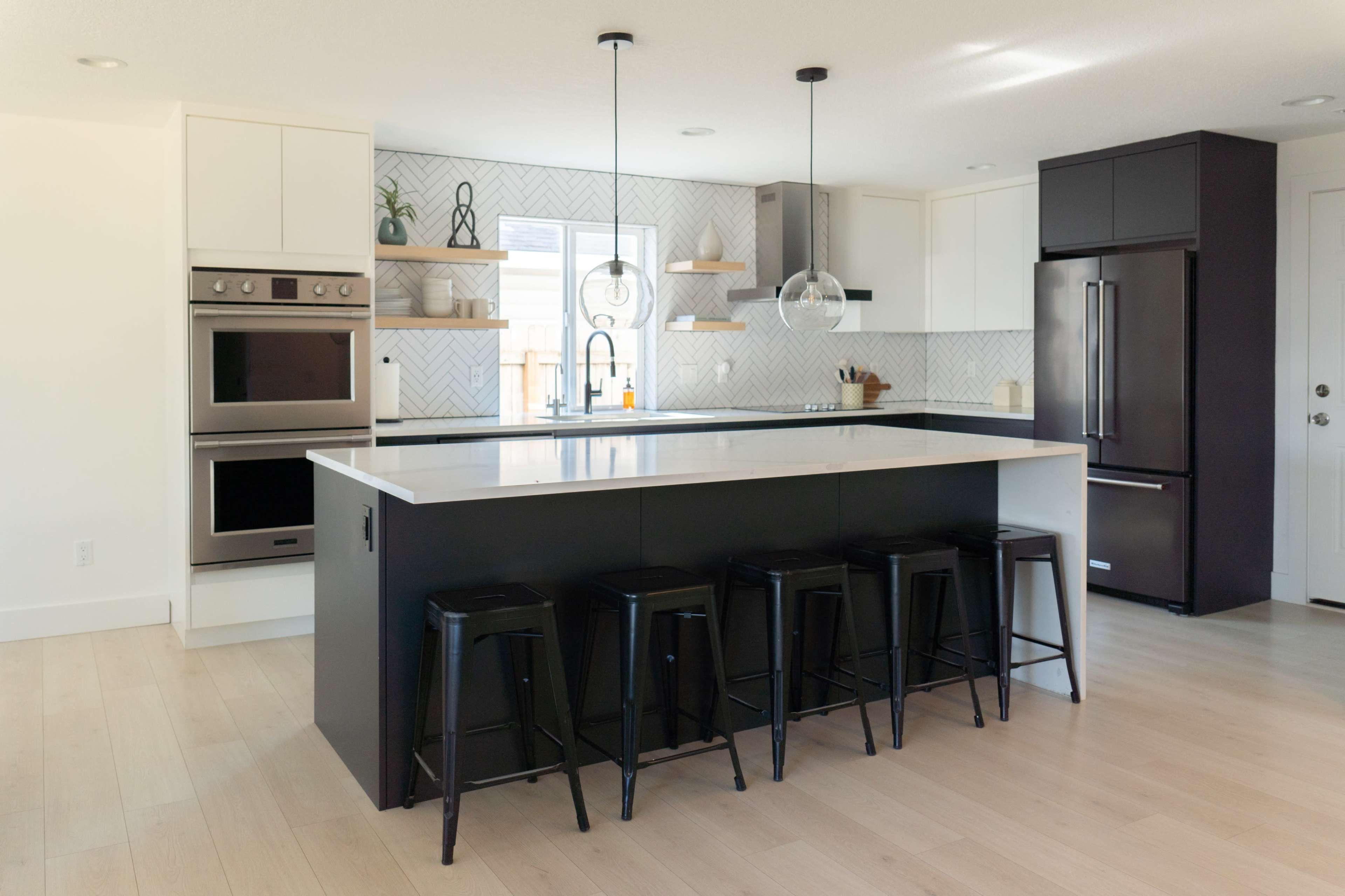 A modern kitchen features a large island with black stools, stainless steel appliances, and light wood flooring.