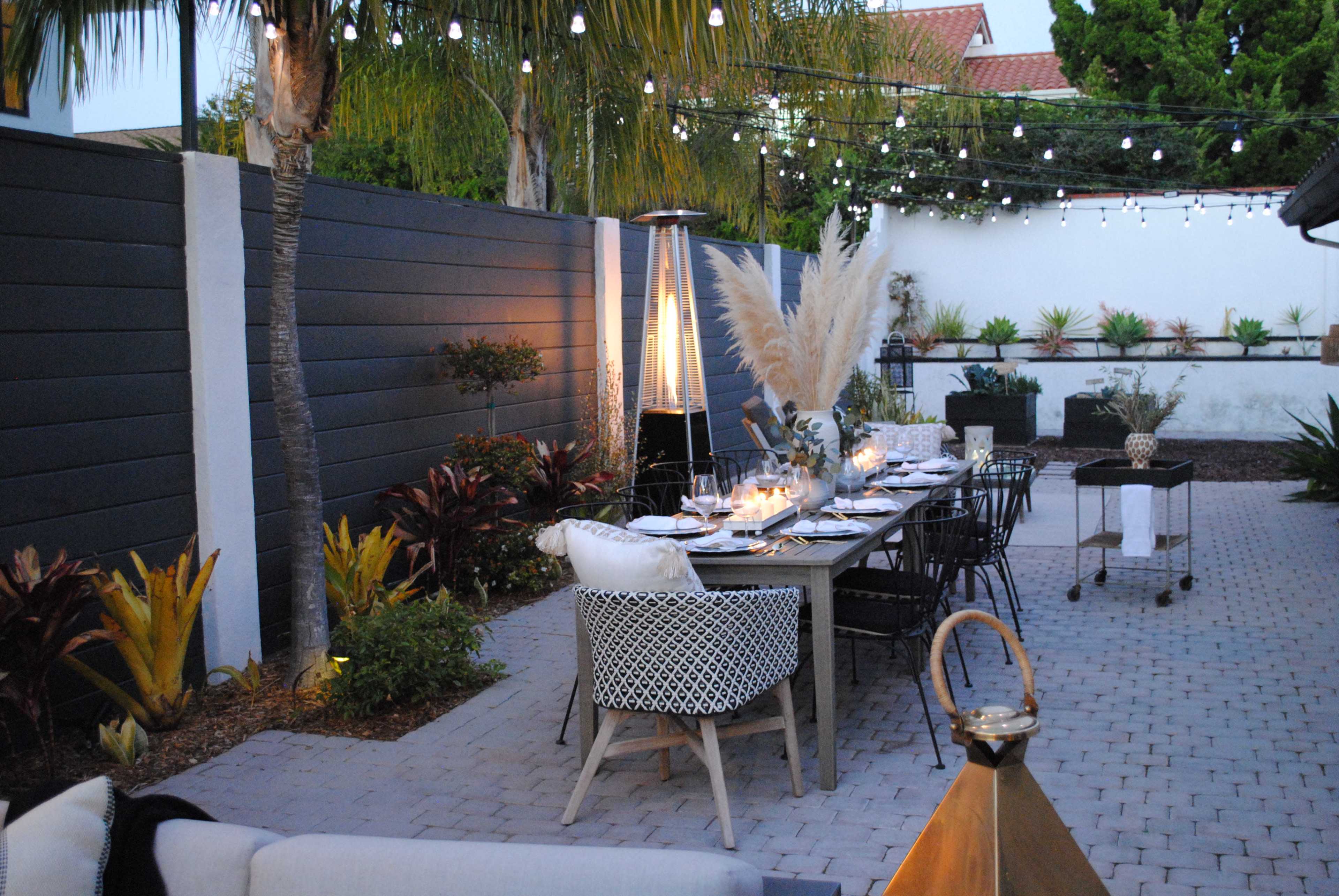 An outdoor dining area with a long table set for a meal, flanked by decorative plants and illuminated by string lights.