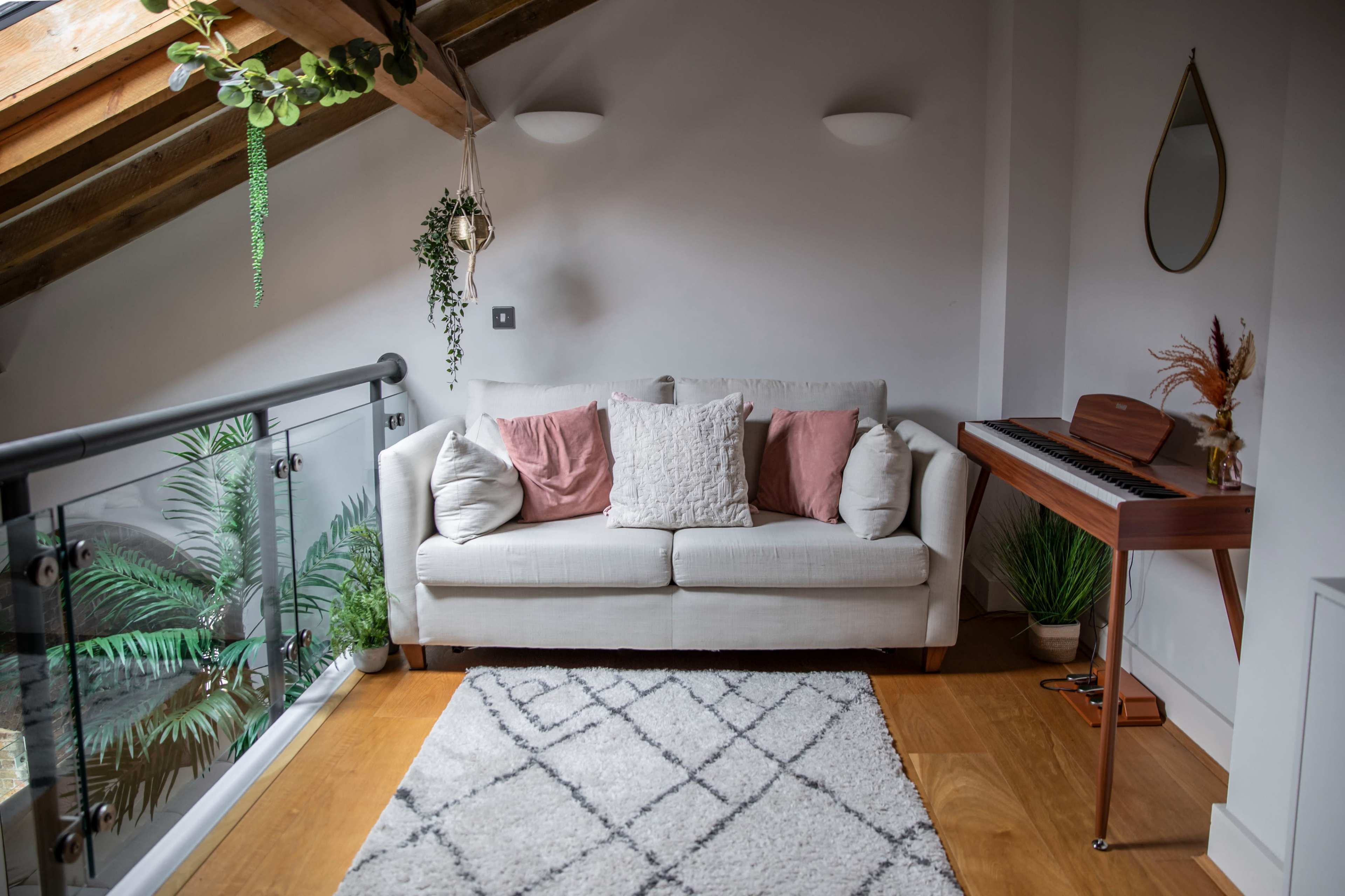 A light-filled living space features a sofa with throw pillows, a keyboard, and plants next to a balcony with a glass railing.