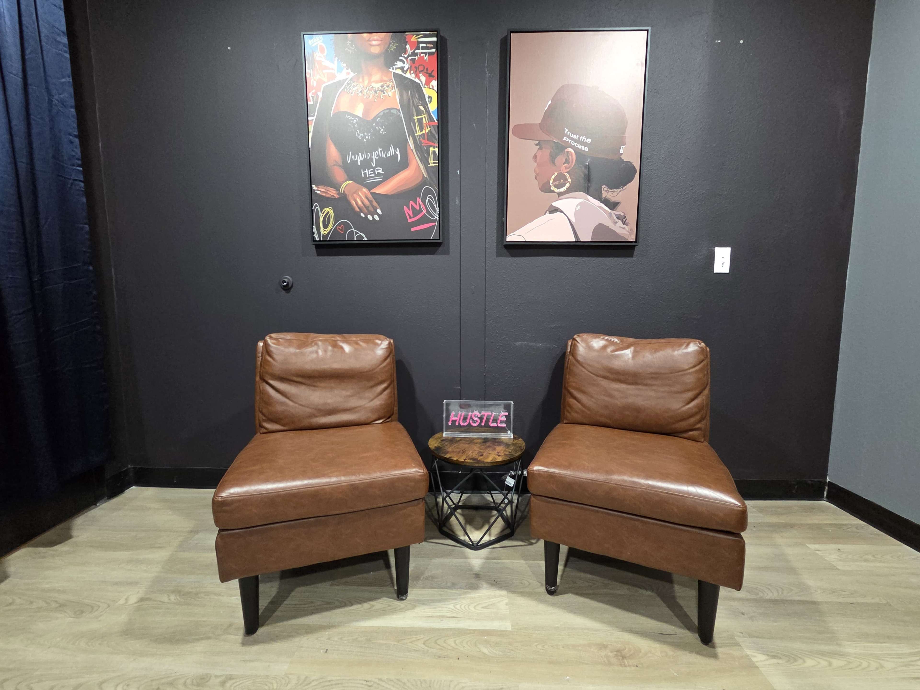 The image shows a cozy seating area with two brown armchairs flanking a round side table, against a dark wall adorned with two framed art pieces.