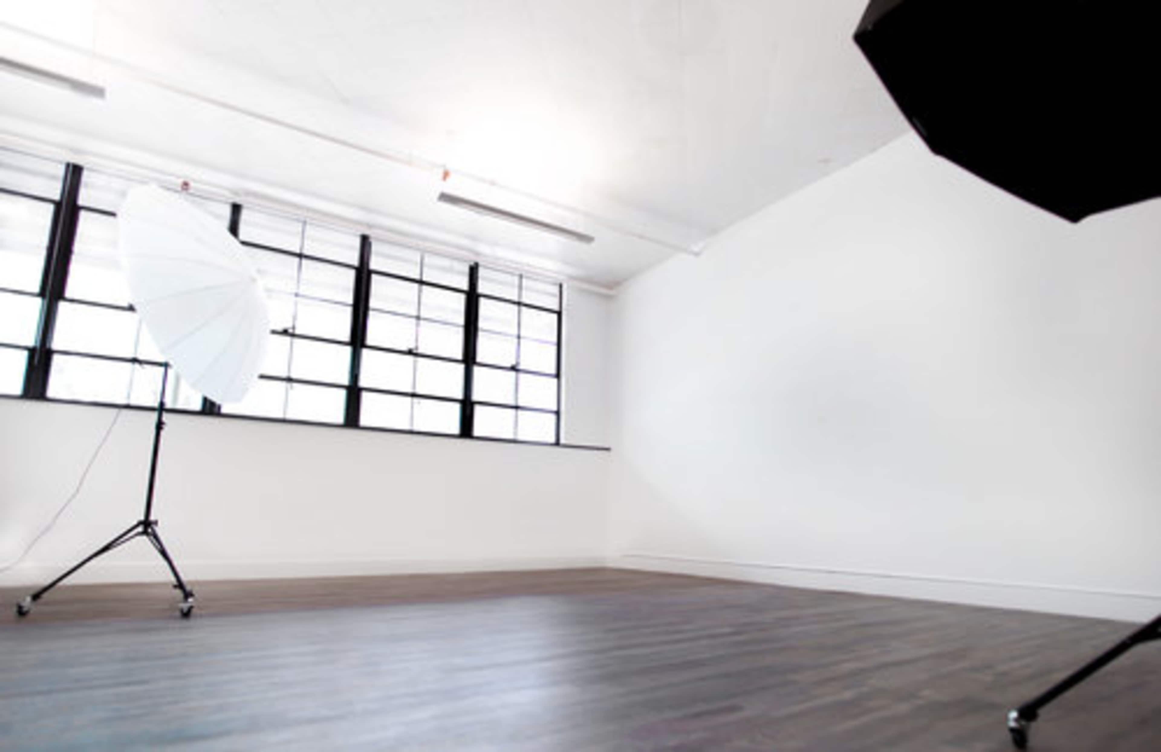 A well-lit photography studio features a large umbrella softbox and a spacious wooden floor.