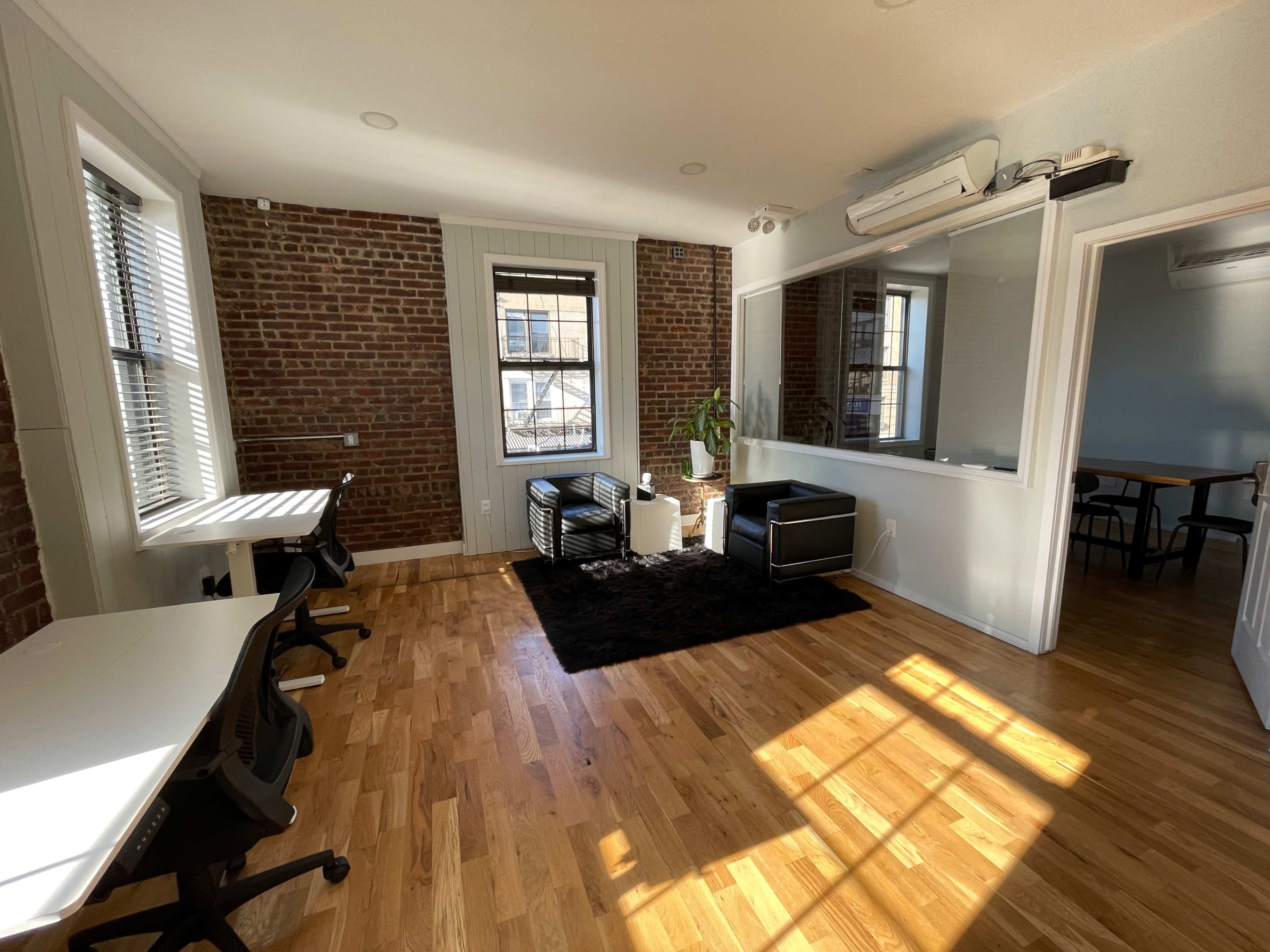 A bright office space with hardwood flooring, exposed brick walls, two black chairs, a small table, a plant, and windows allowing natural light to enter.