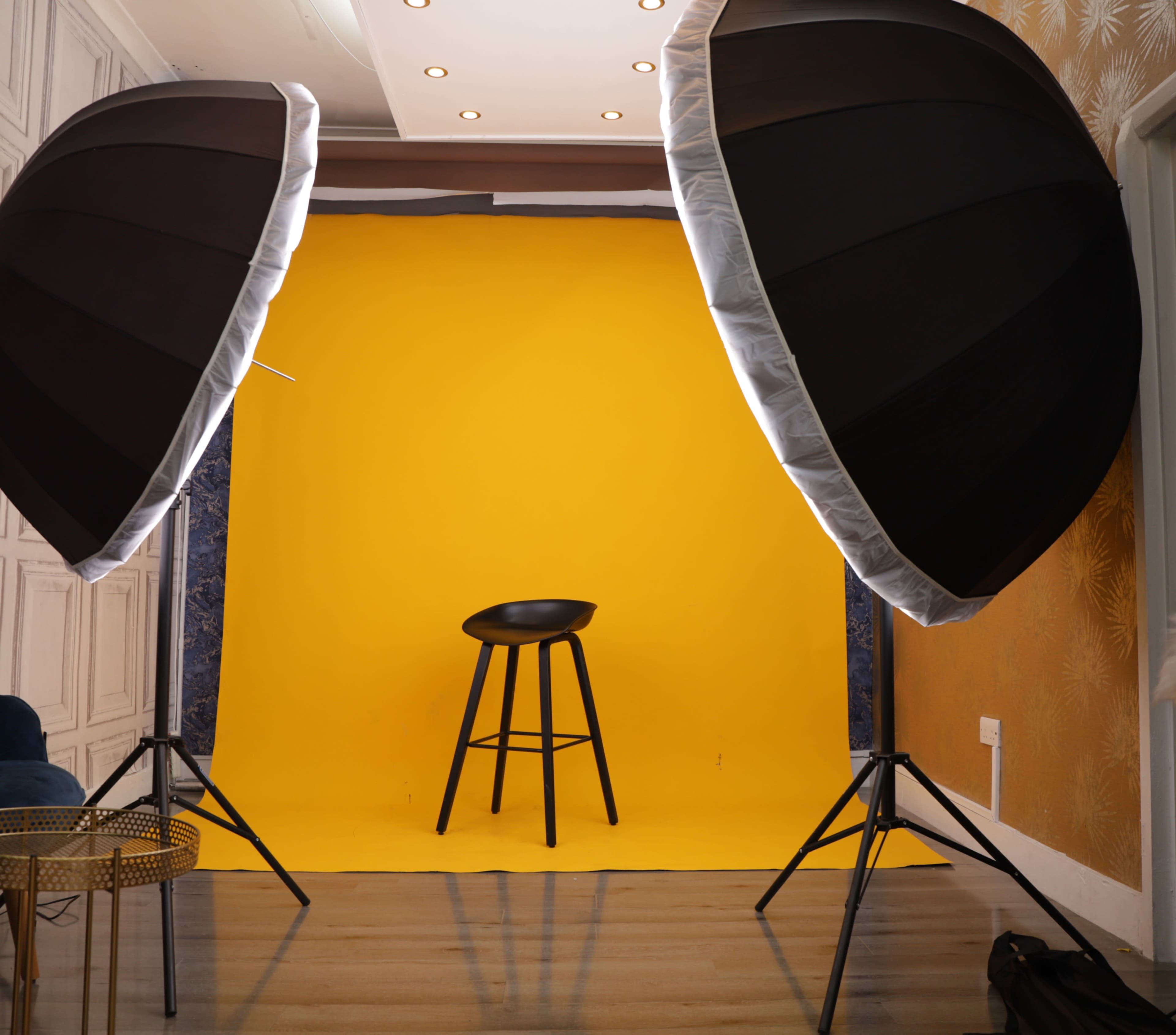 A photography studio setup features two large softbox lights flanking a black stool in front of a bright yellow backdrop.
