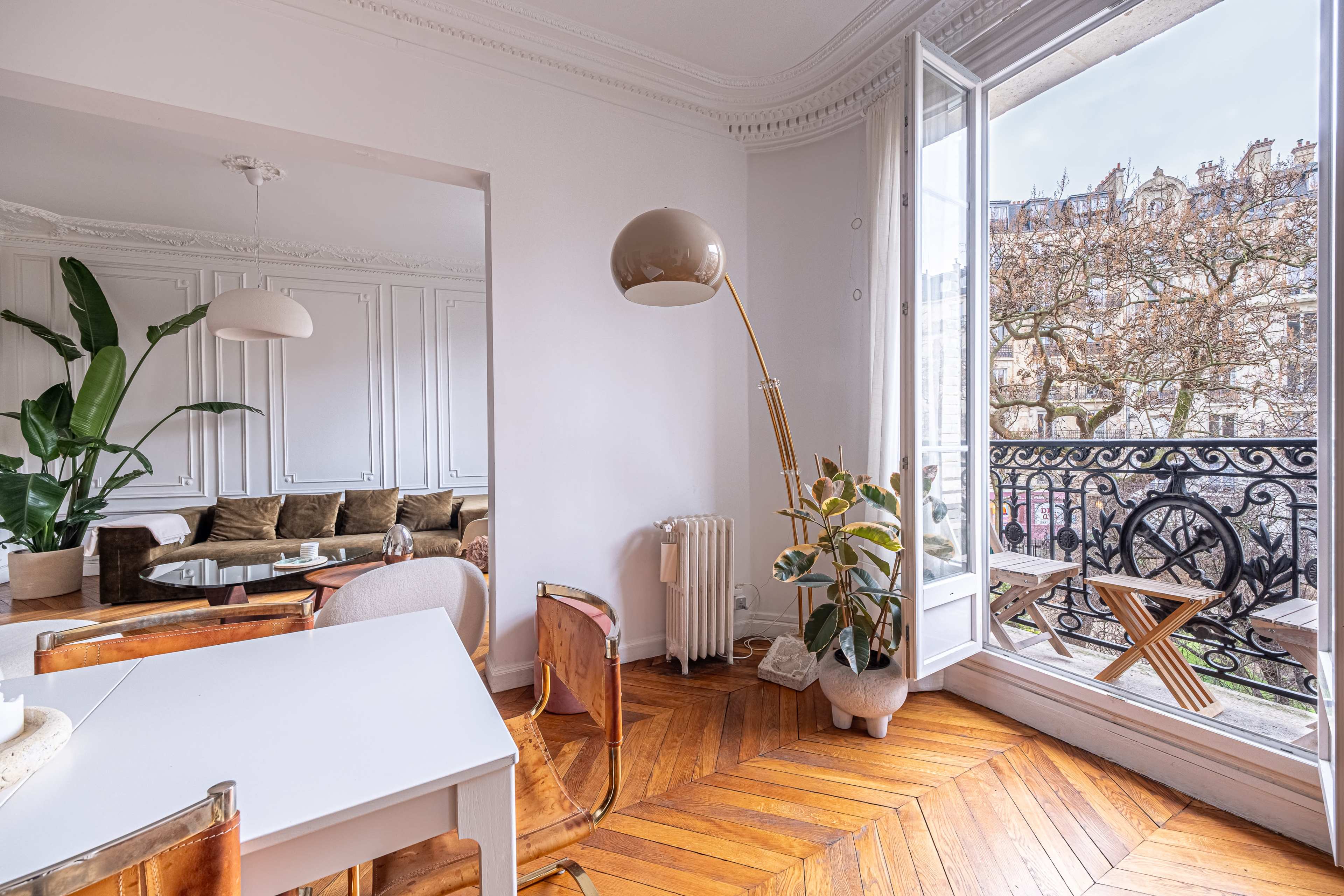 The image shows a bright, modern apartment with a spacious living area featuring a large window that opens to a balcony overlooking a street with trees.