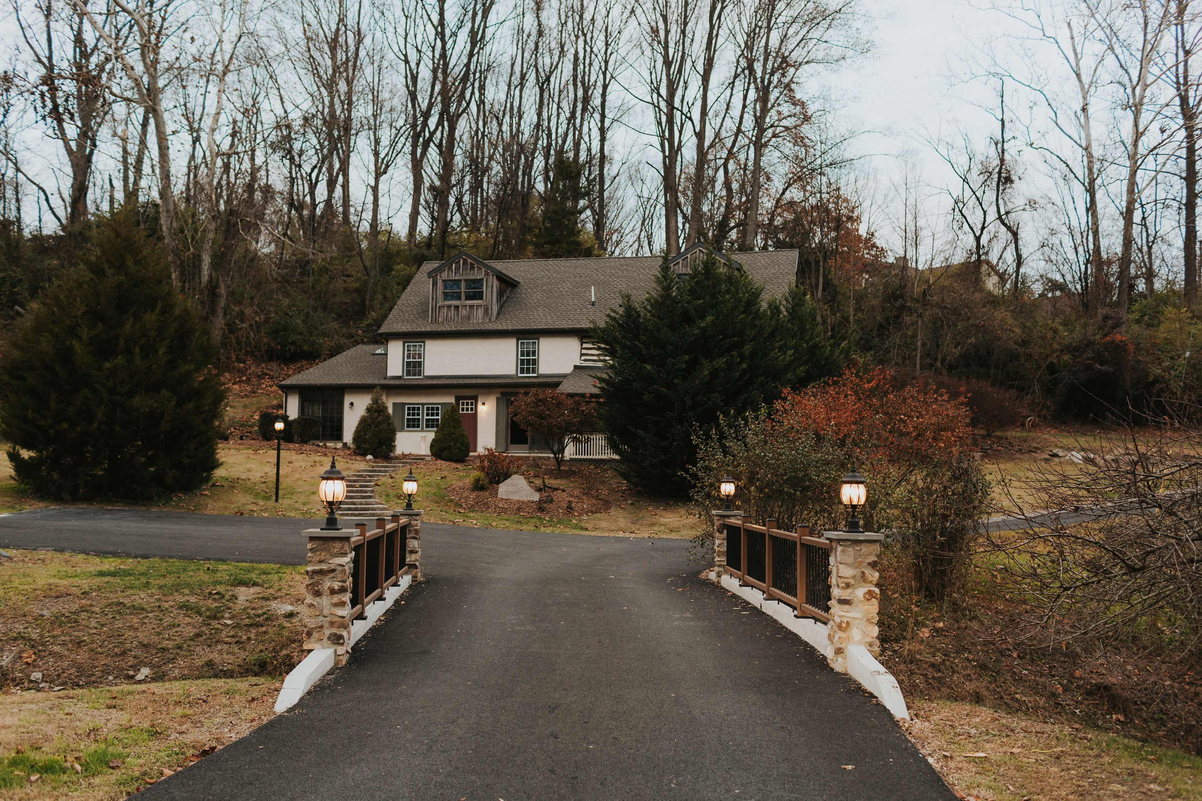 A two-story house with a sloped roof is situated at the end of a tree-lined driveway, leading up to a small bridge.